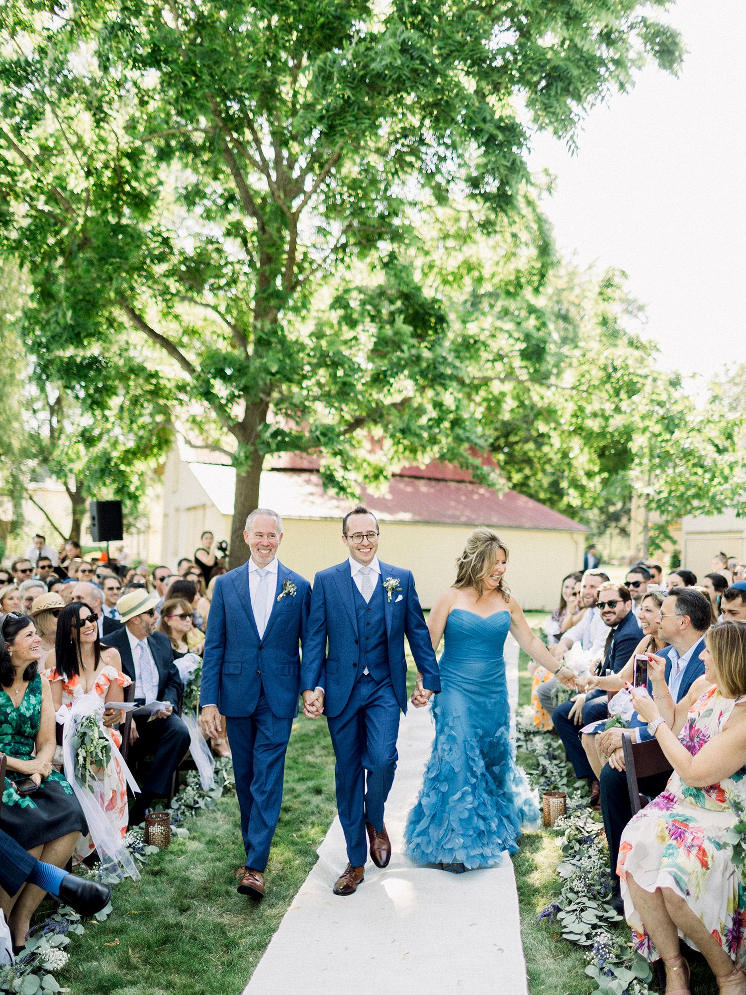 Wedding party walks down outdoor aisle in blue attire while guests watch from both sides.