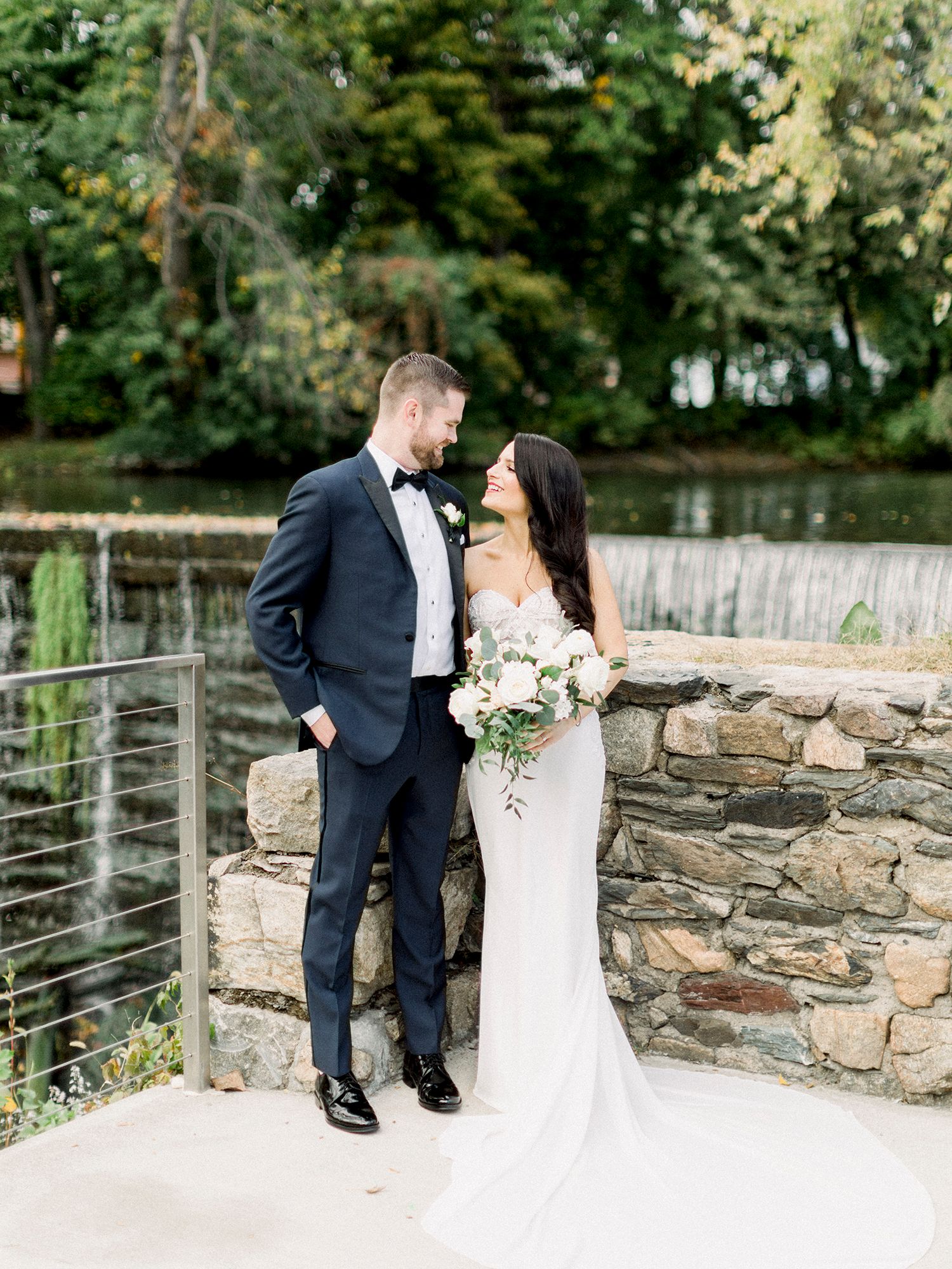 Elegant couple in formal wedding attire posing by stone wall with modern railing.