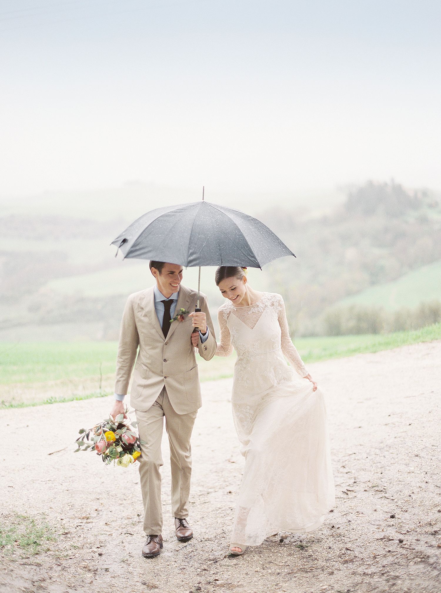 Couple walking with umbrella across misty field in romantic wedding attire.