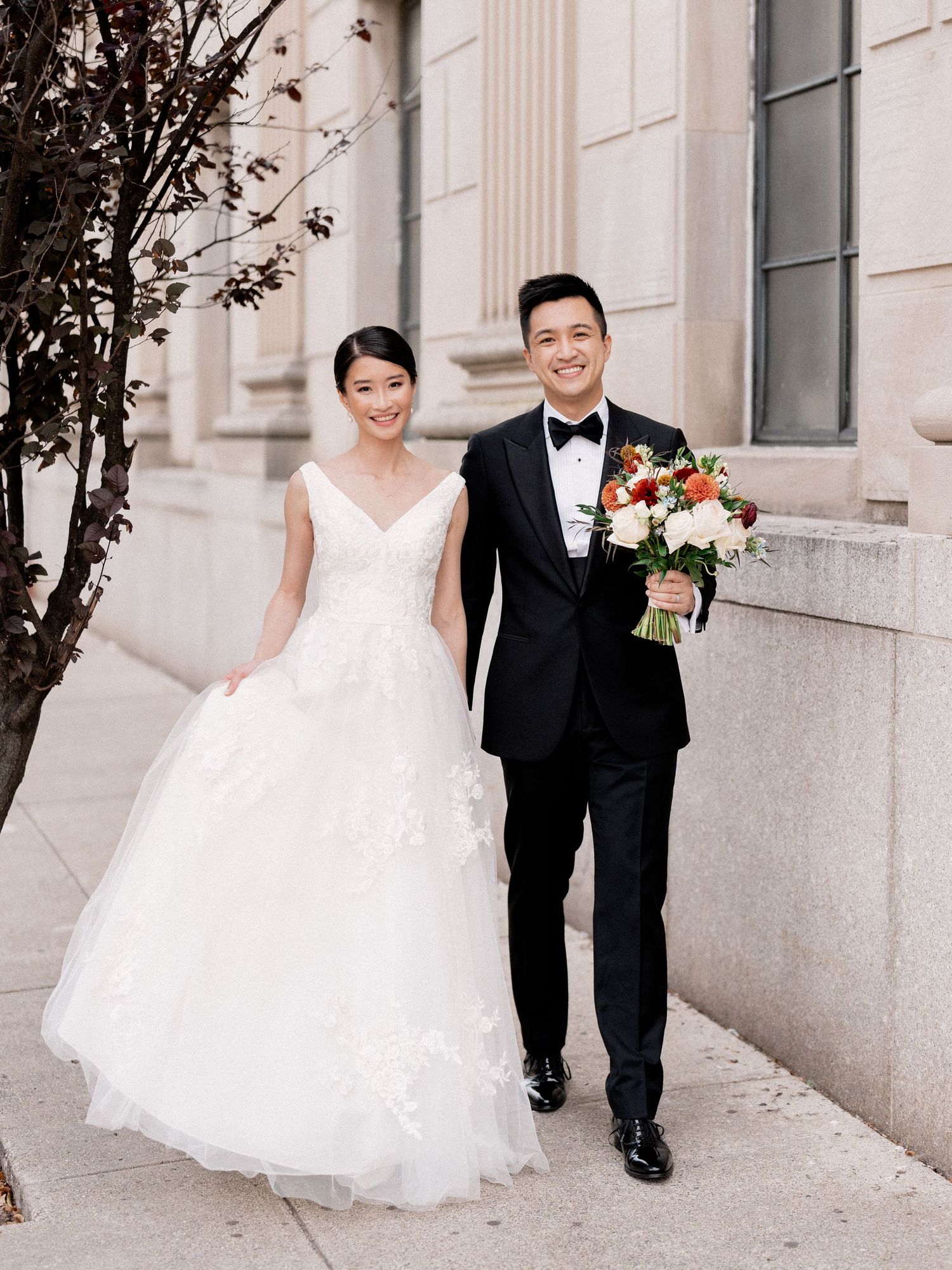 Couple walking together outside a classic stone building with autumn trees.