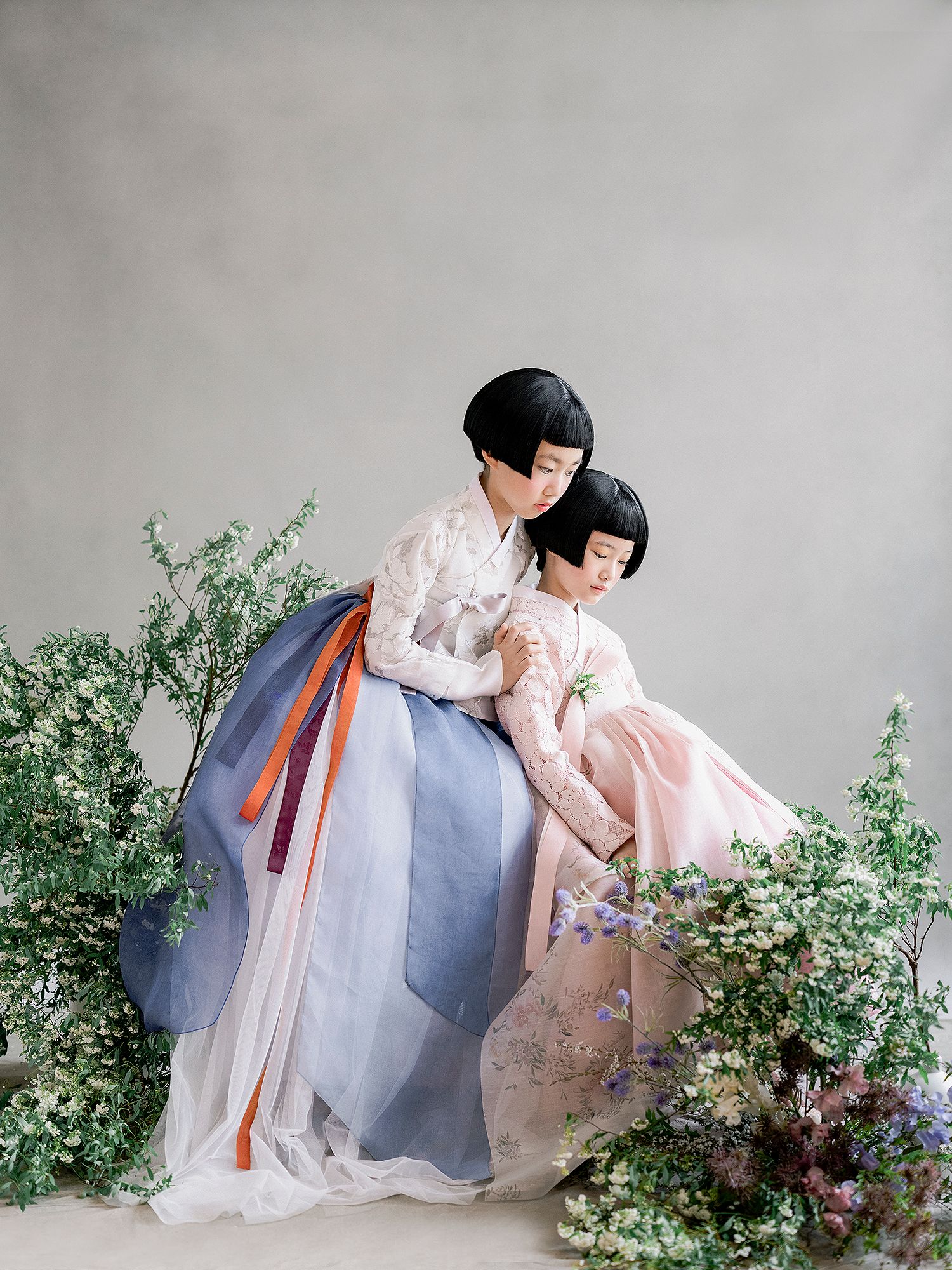 Two people wearing traditional Korean hanbok dresses pose elegantly among delicate floral arrangements against a gray backdrop.