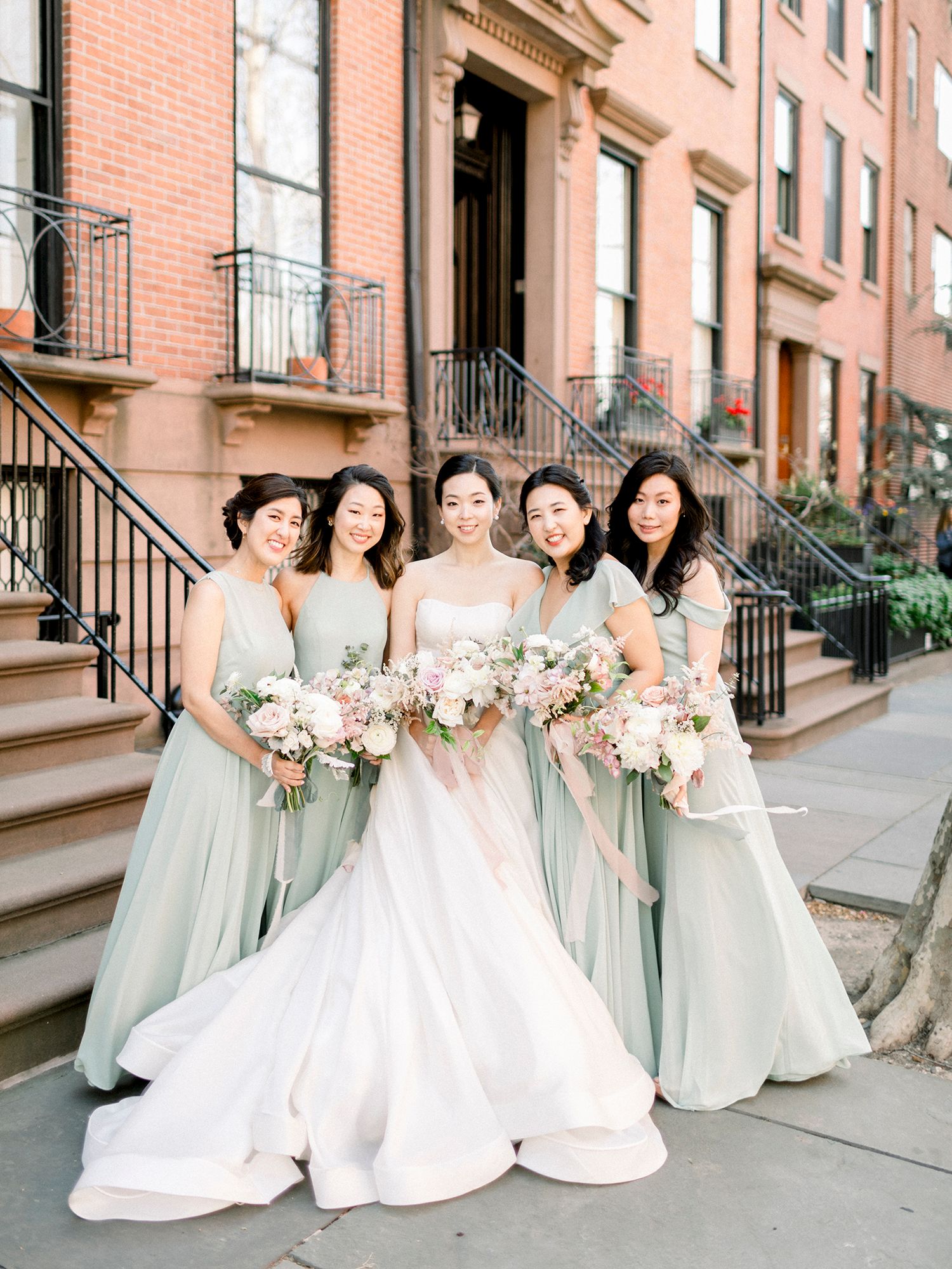 Bridesmaids in mint green dresses pose with bride in white gown on brownstone steps.