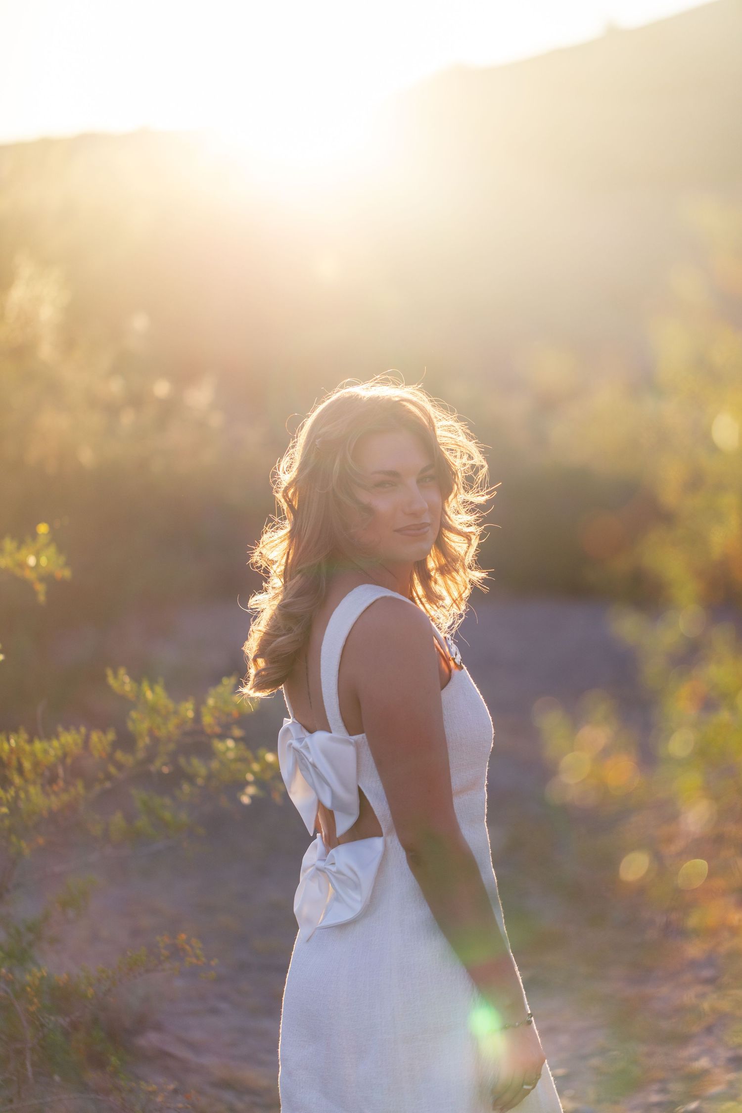 Woman in white backless dress standing in golden sunlight with bokeh nature background.