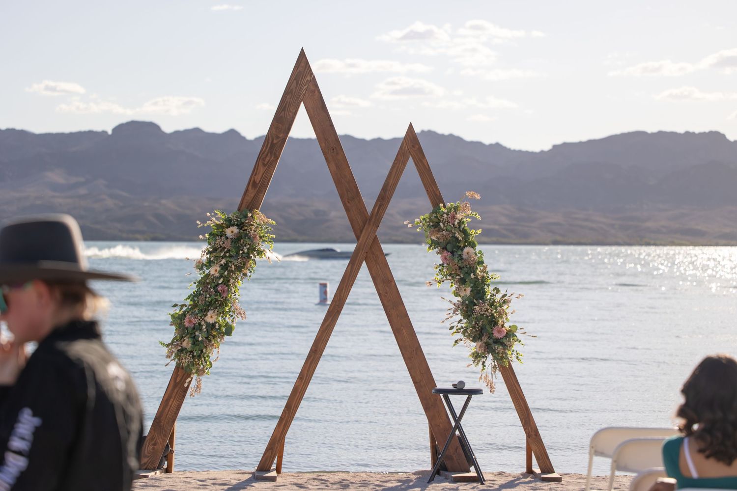 Rustic wooden triangular wedding arch decorated with greenery stands on beach overlooking mountain lake view in Lake Havasu City Arizona