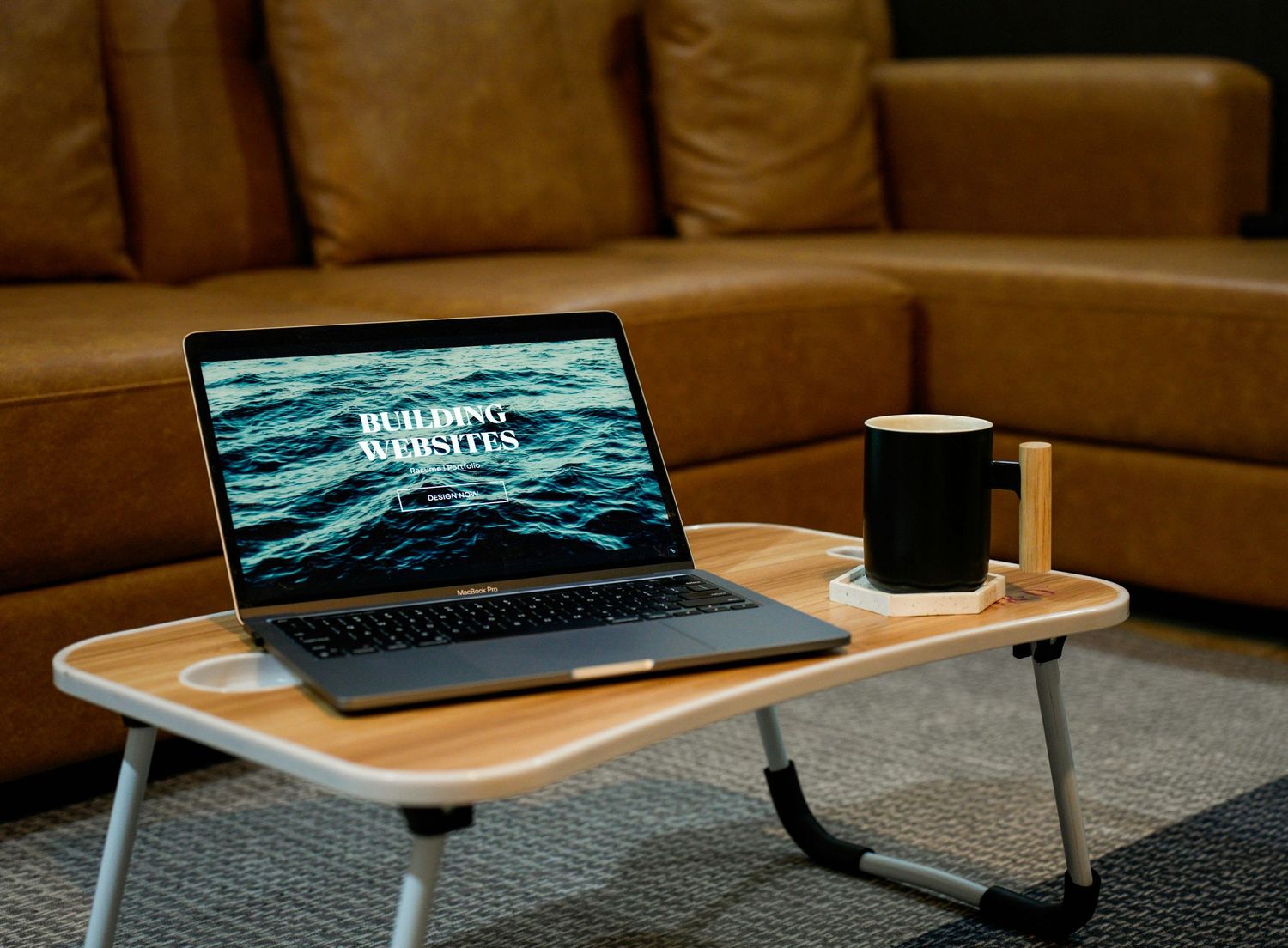 Laptop displaying ocean waves screensaver with coffee cup on wooden lap desk near leather couch.