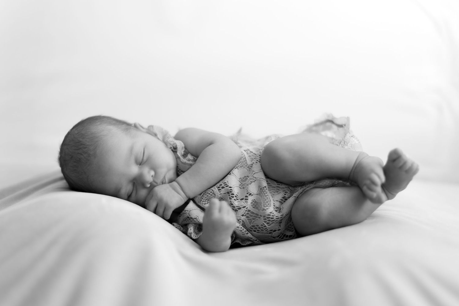 Black and white newborn baby sleeping peacefully on soft white bedding wearing a delicate lace romper.