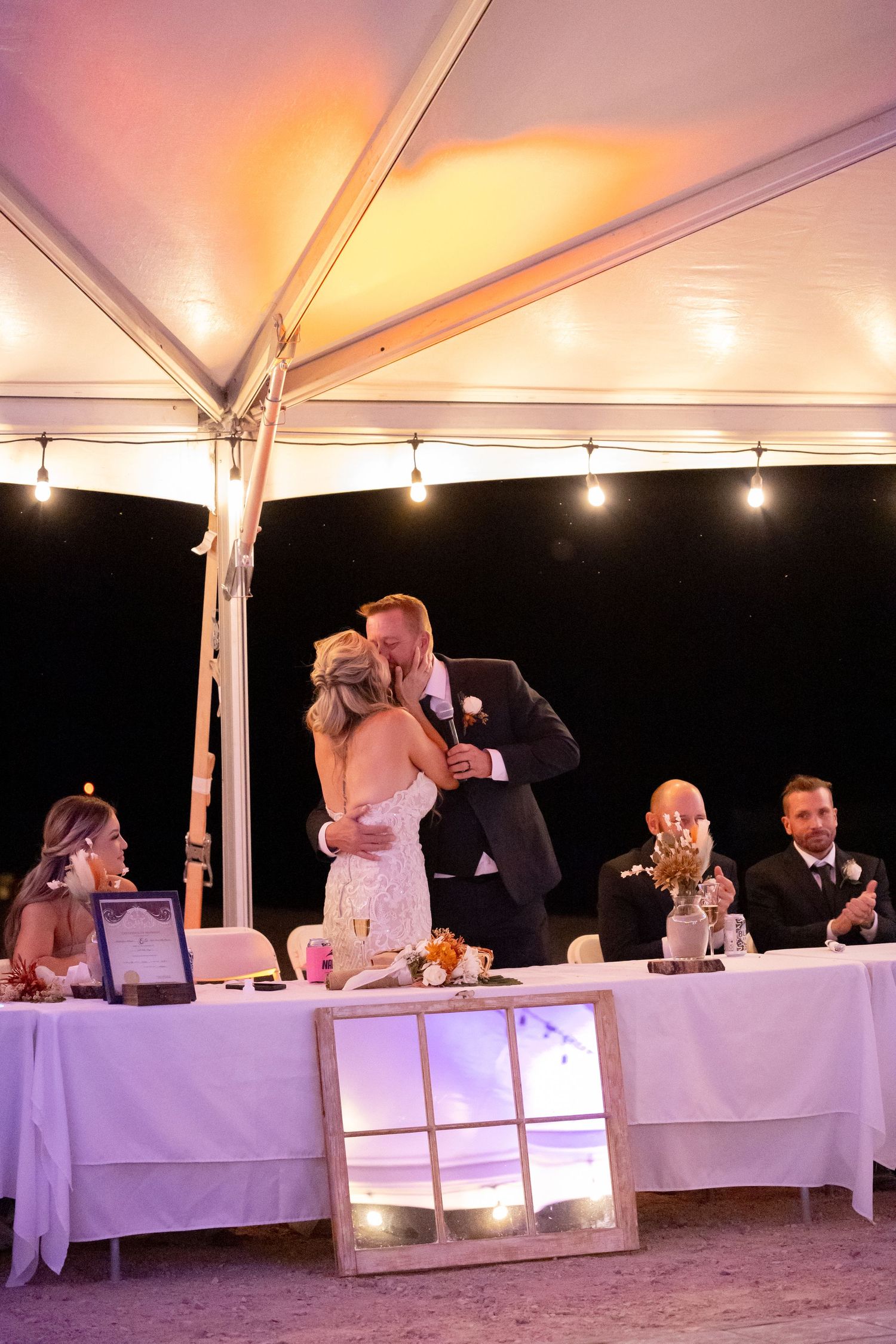A romantic couple share a kiss under an illuminated outdoor tent during their wedding reception with guests seated in Lake Havasu City AZ