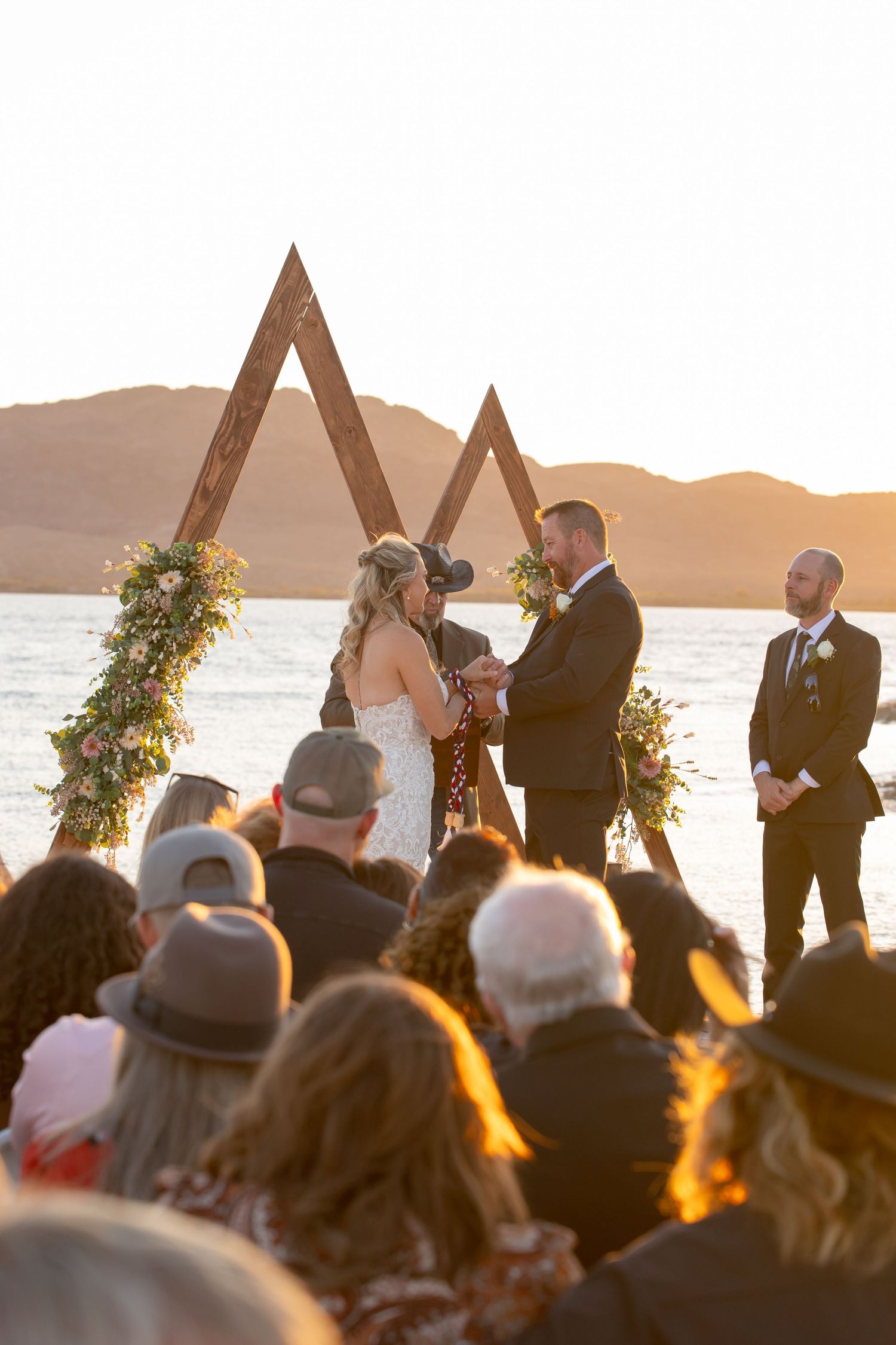 A scenic lakeside wedding ceremony takes place at sunset under triangular wooden arches decorated with greenery at the riviera lake havasu