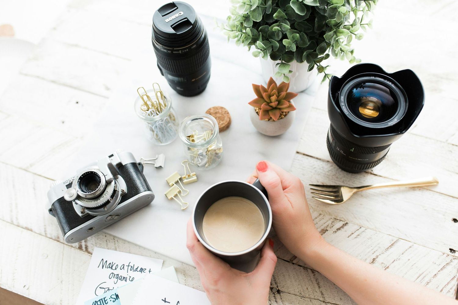 Flatlay of photography equipment including camera lenses, coffee cup and desk accessories.