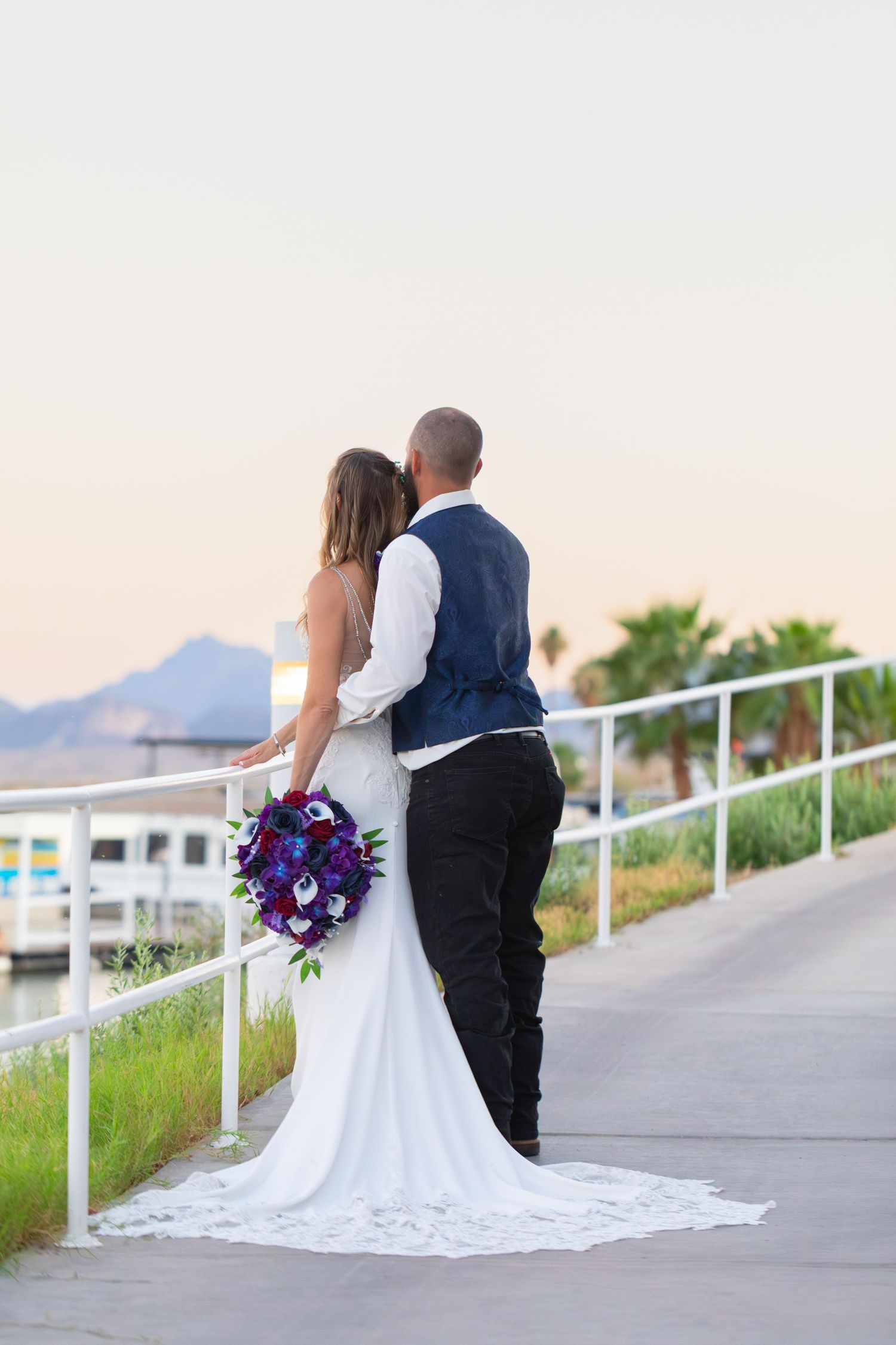 Bride and groom stand together on a waterfront promenade at sunset, bride holding a vibrant purple bouquet.