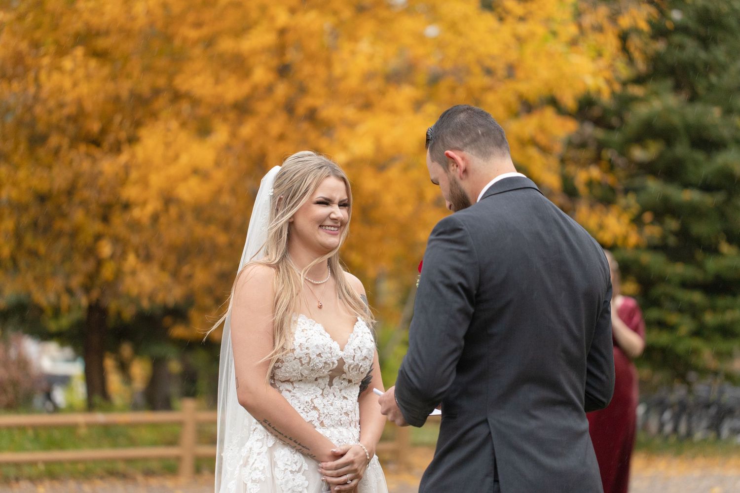 Smiling bride in lace gown and veil holds hands with groom in gray suit outdoors with fall foliage backdrop.