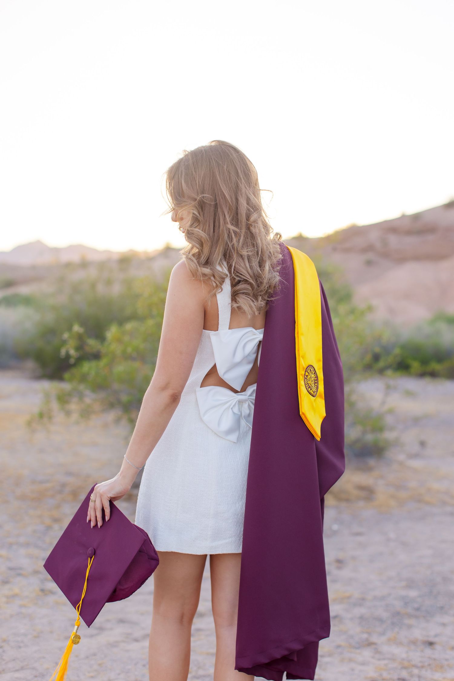 Young woman in white dress holds maroon graduation cap and gown with gold stole in desert landscape at sunset.