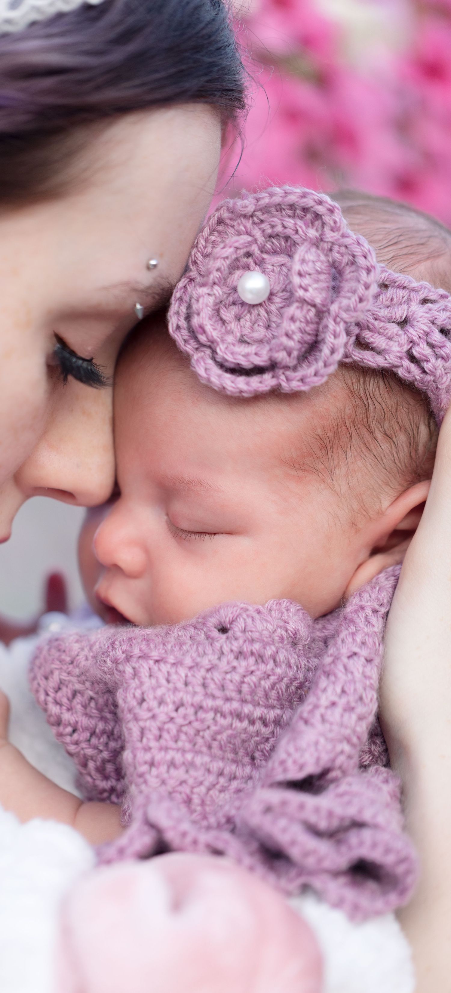 Mother tenderly nuzzles sleeping newborn baby wearing purple crochet flower headband and matching outfit.
