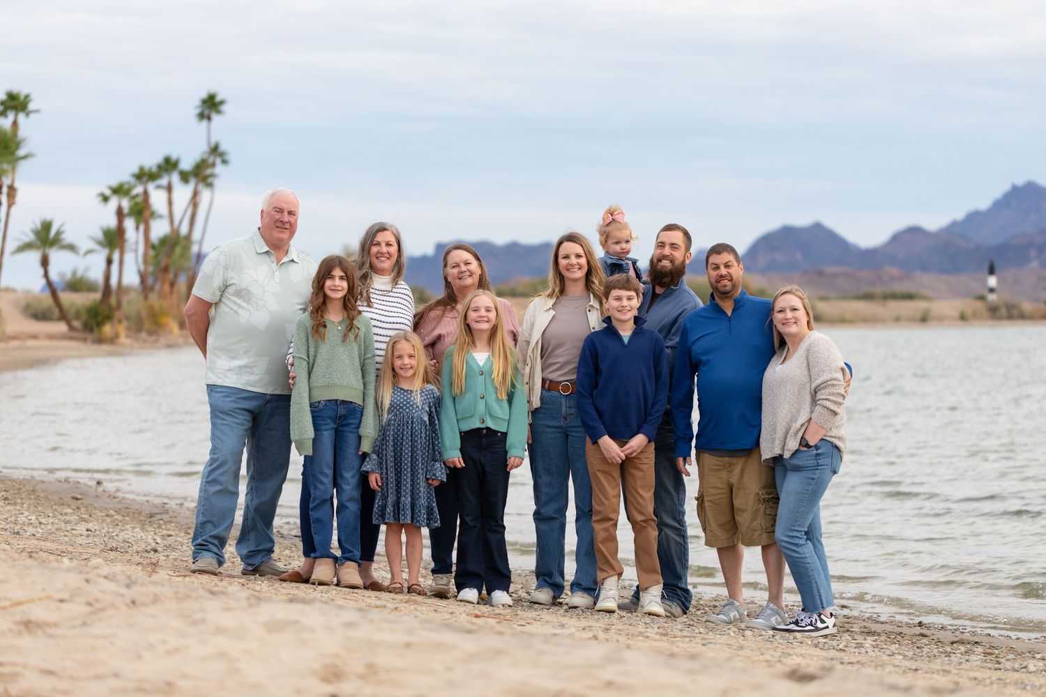 Large family group smiling together on a sandy beach with mountains and palm trees in the background.