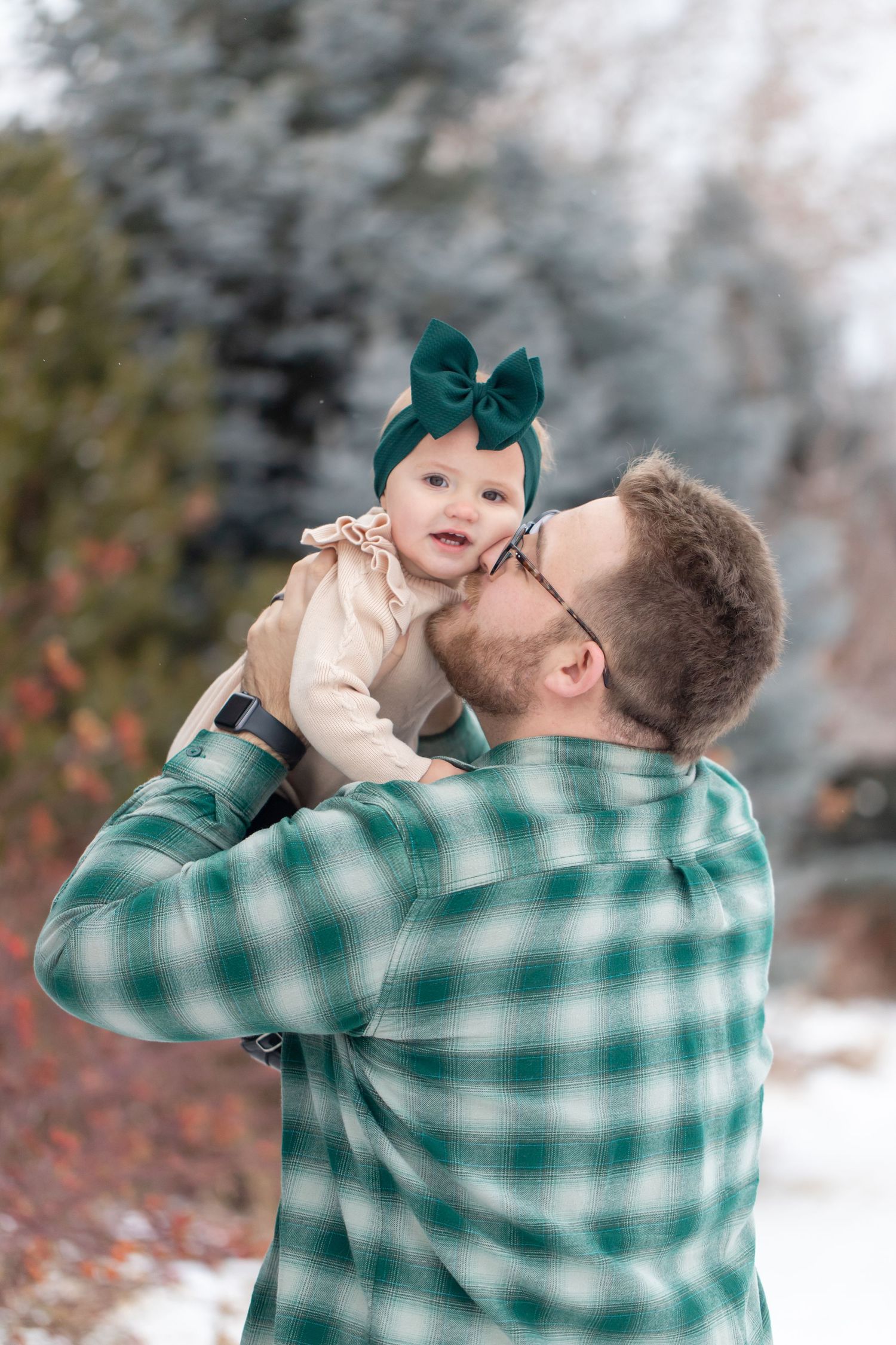 A person in a green plaid shirt holds a baby wearing a teal bow in a snowy winter setting.
