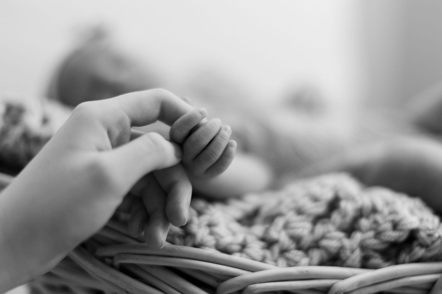 Black and white photo of a newborn baby holding an adult finger, lying on a knit blanket in a basket.