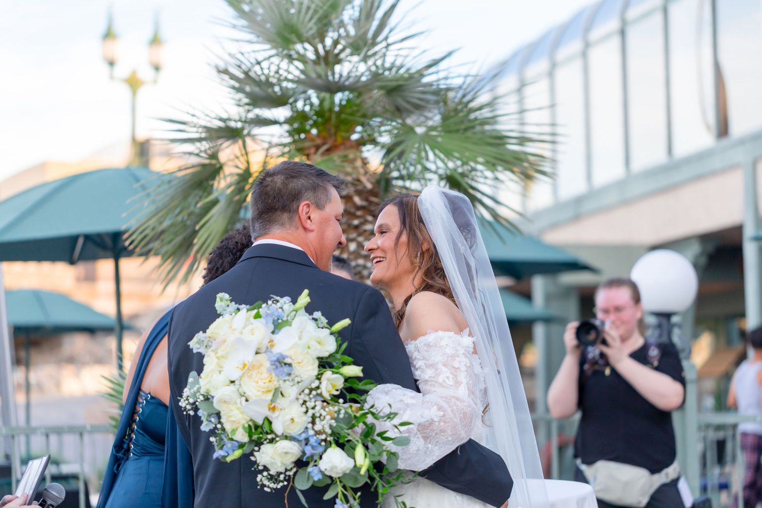 Bride and groom share first kiss at outdoor wedding ceremony with palm tree backdrop and photographer capturing the moment at Shugrues