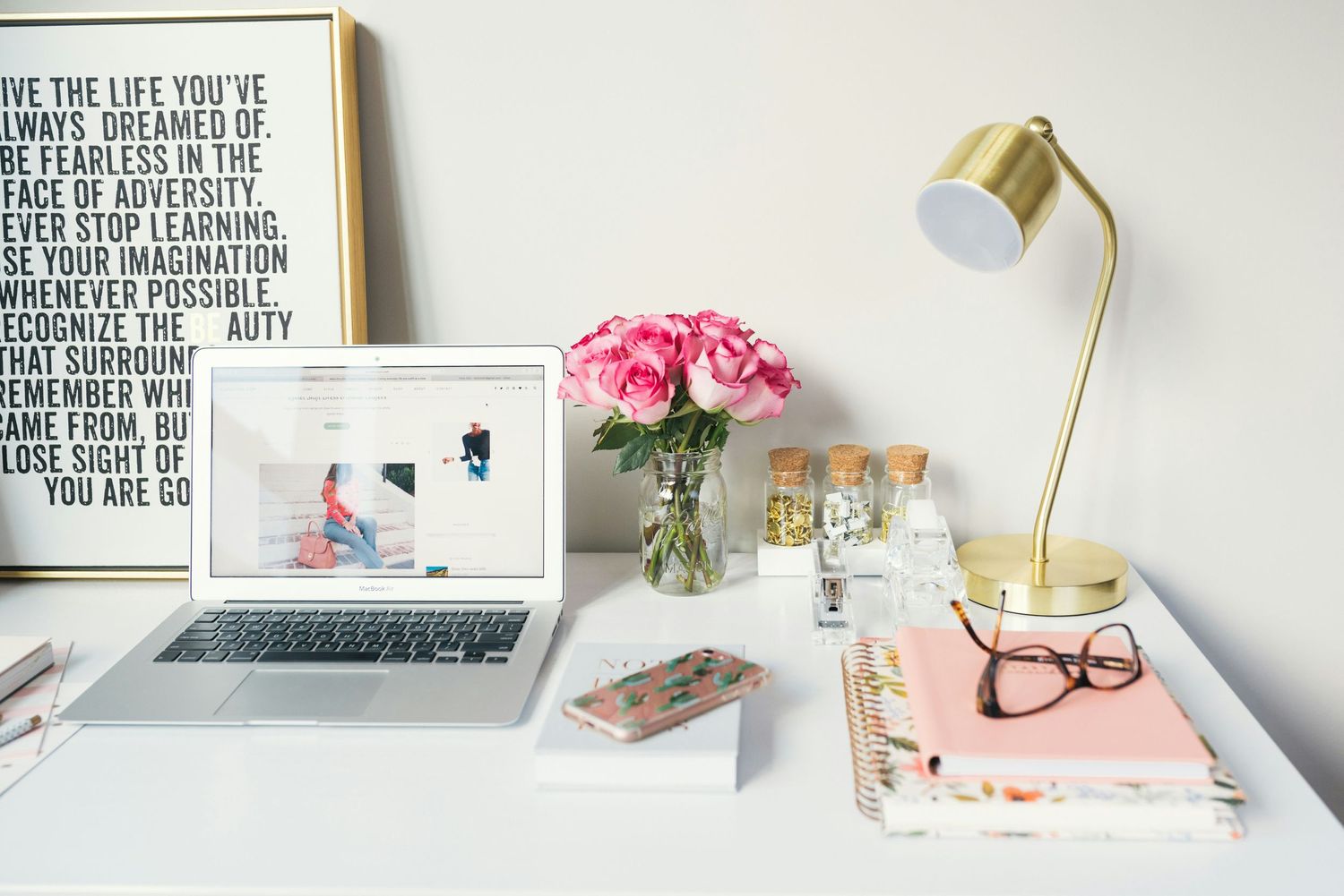 Modern workspace flatlay with laptop, pink roses in vase, gold desk lamp, and pink stationery on white desk.
