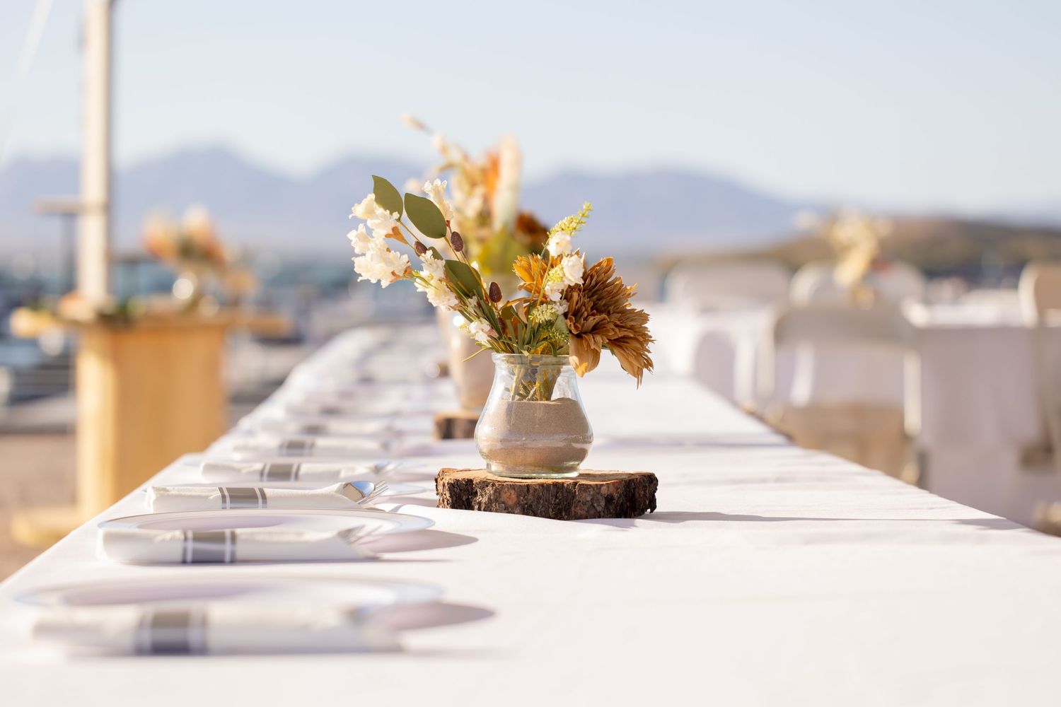 White table setting with rustic floral centerpiece on wood slice at outdoor event with mountain views in background in Lake Havasu City