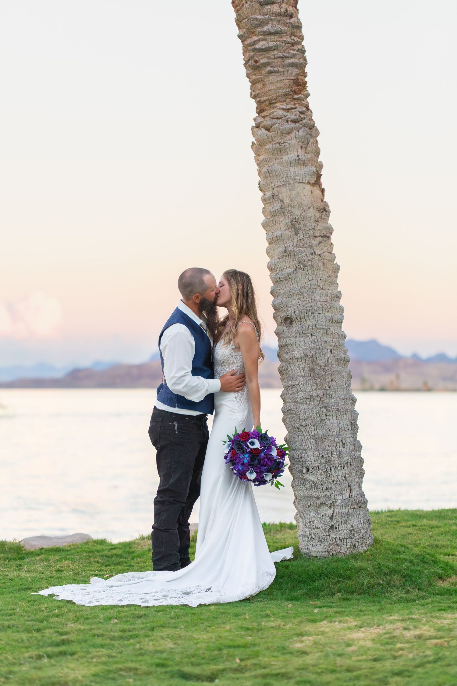 Bride and groom share a kiss near a palm tree by a lakeside at sunset, bride holding purple bouquet at the Nautical Resort in Lake Havasu