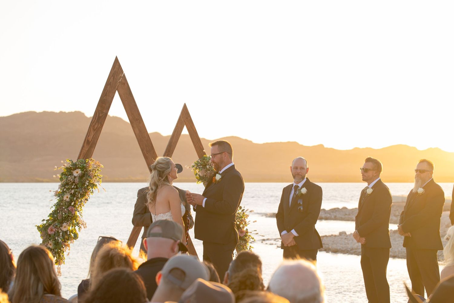 A scenic lakeside wedding ceremony at sunset with a triangular wooden arch decorated with greenery and flowers at the riviera lake havasu