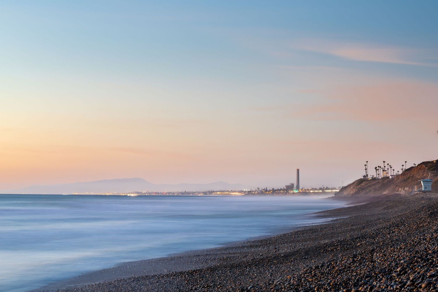Coastal california landscape at sunset with dark rocky beach and calm ocean waters stretching to the horizon.