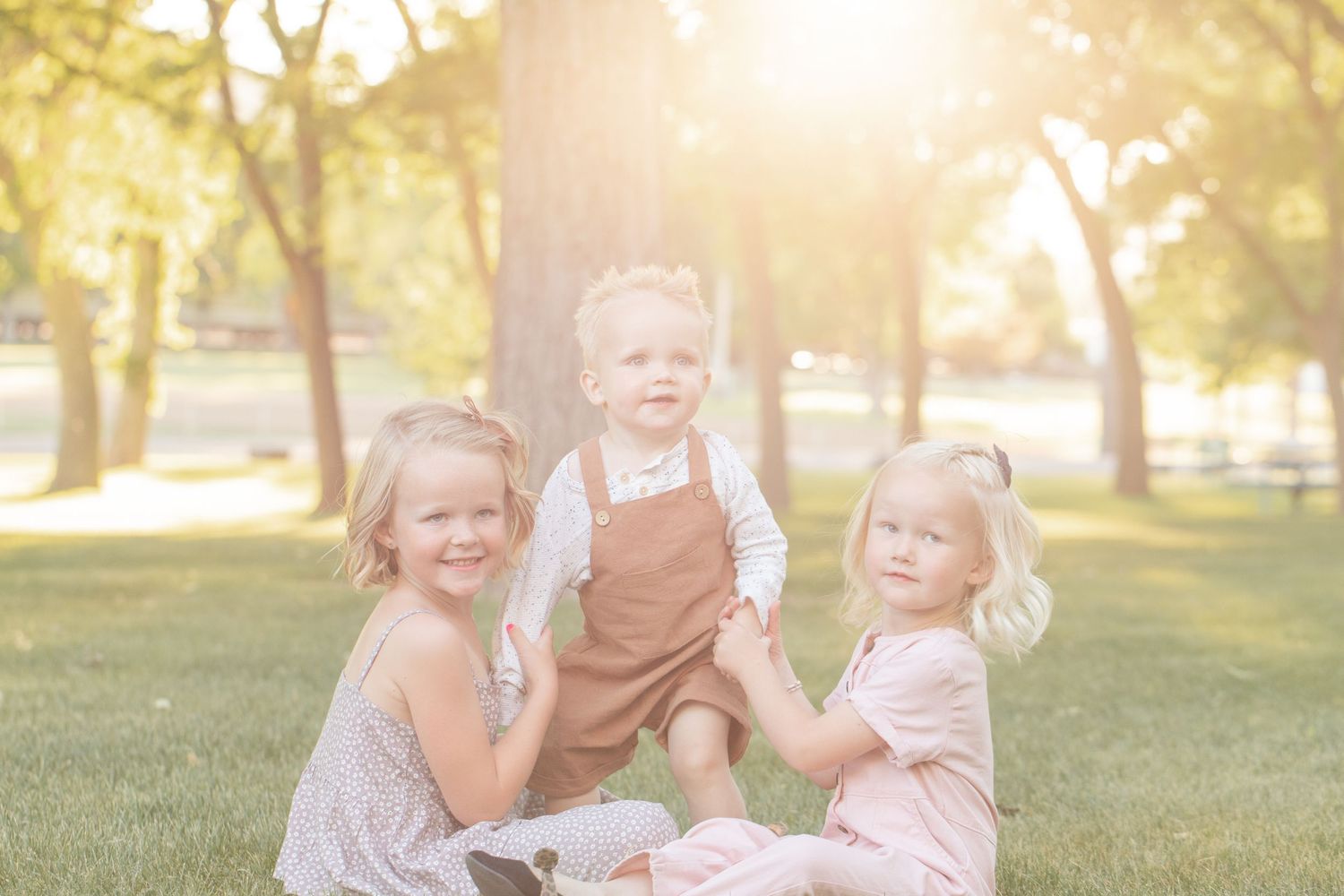 Three young children pose together in a sunlit park, two girls in pink dresses holding a toddler boy in brown overalls.