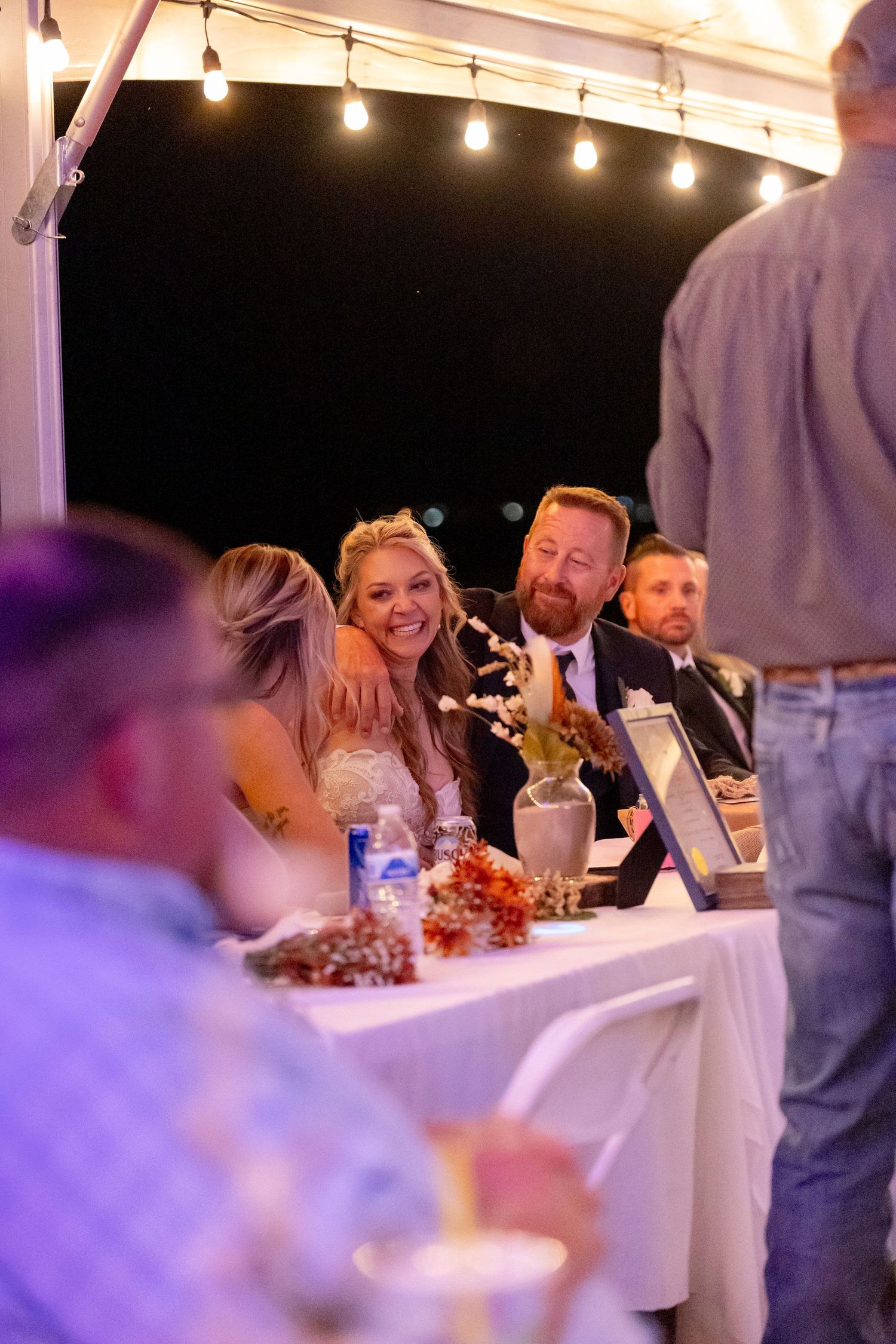 Bride and groom laughing at wedding reception table under string lights at outdoor evening celebration at The Riviera in Lake Havasu City
