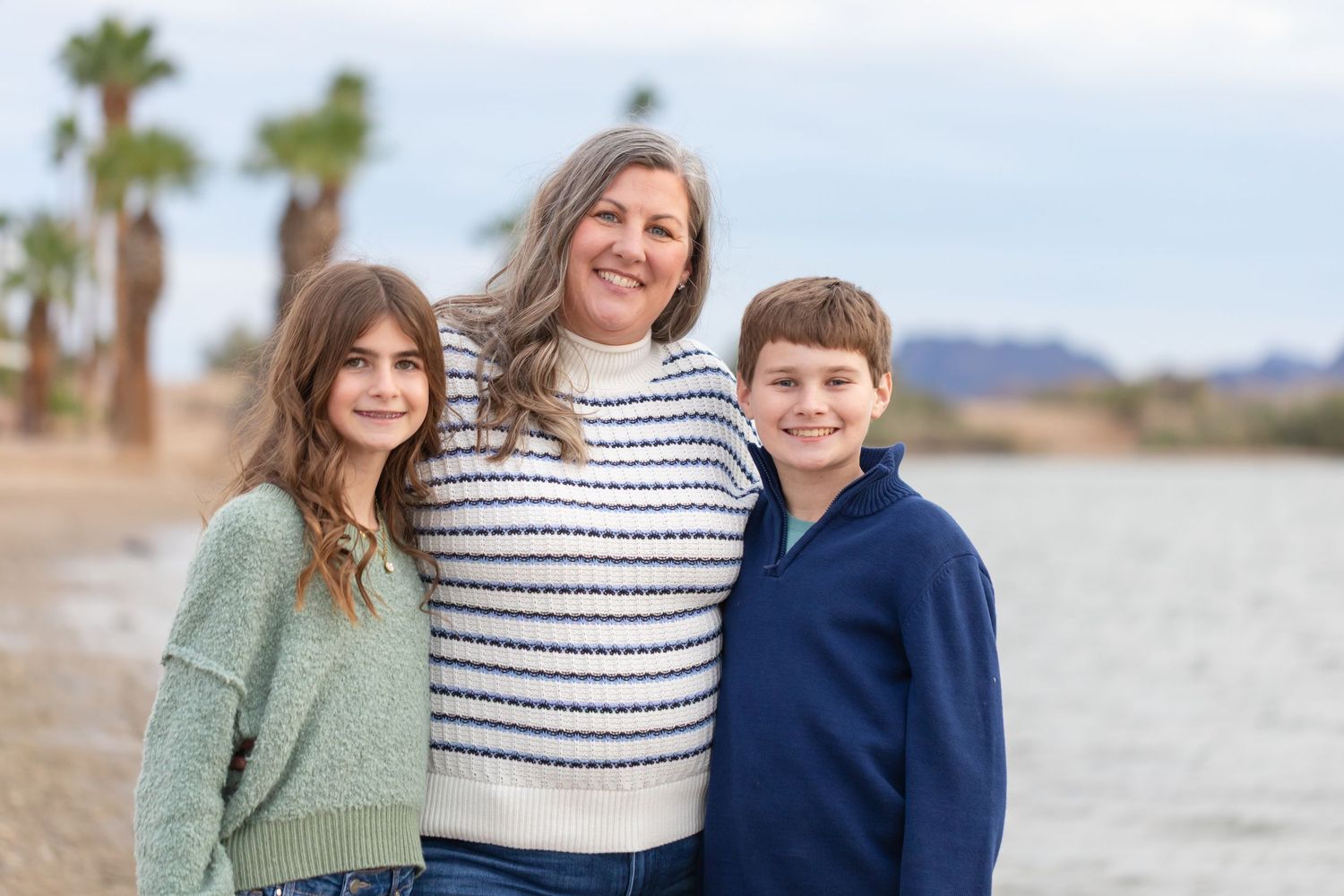 A woman and two children smile together near a lake with palm trees and mountains in the background at rotary park in Lake Havasu City