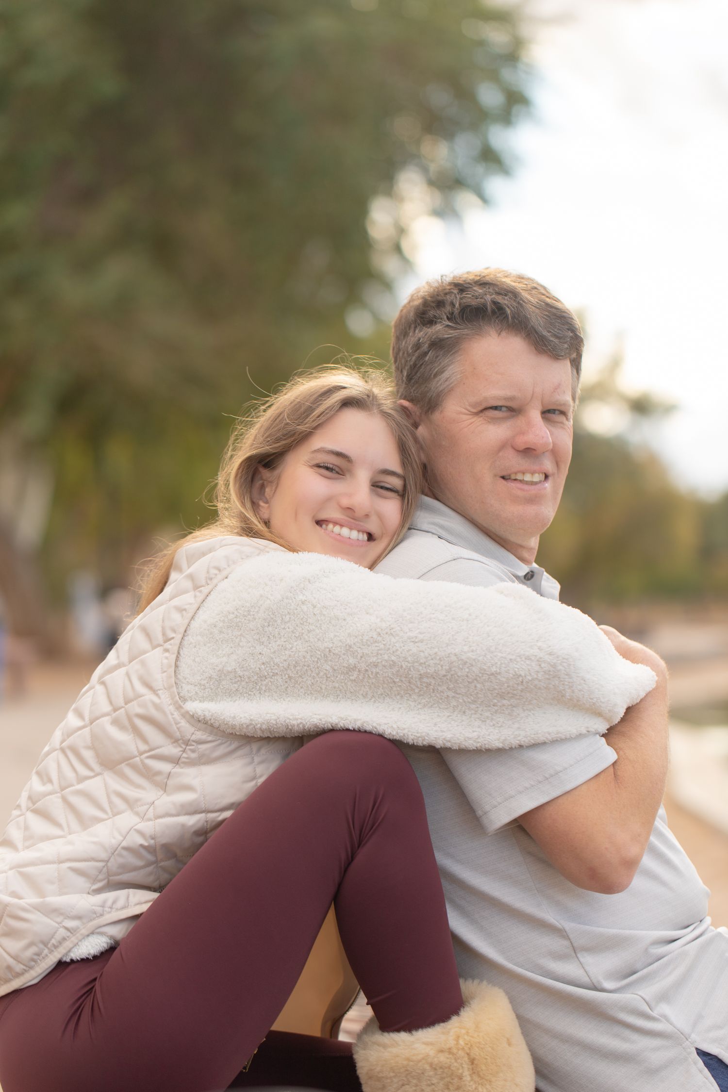A smiling couple embraces outdoors, woman in white vest hugging man from behind near trees at the bridgewater channel in Lake Havasu City