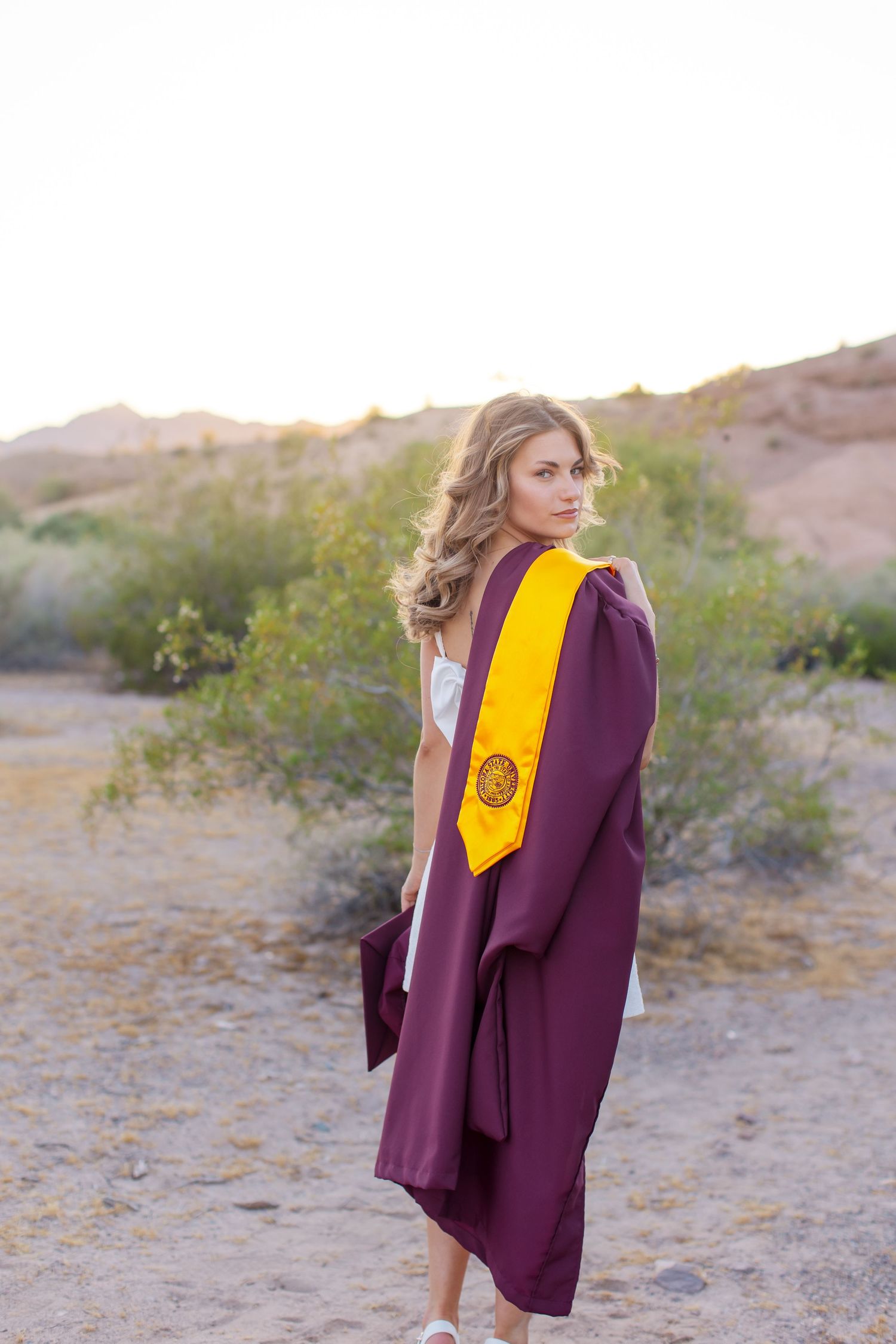Young woman in maroon graduation gown and gold stole poses outdoors in desert landscape at golden hour in Lake Havasu City Arizona