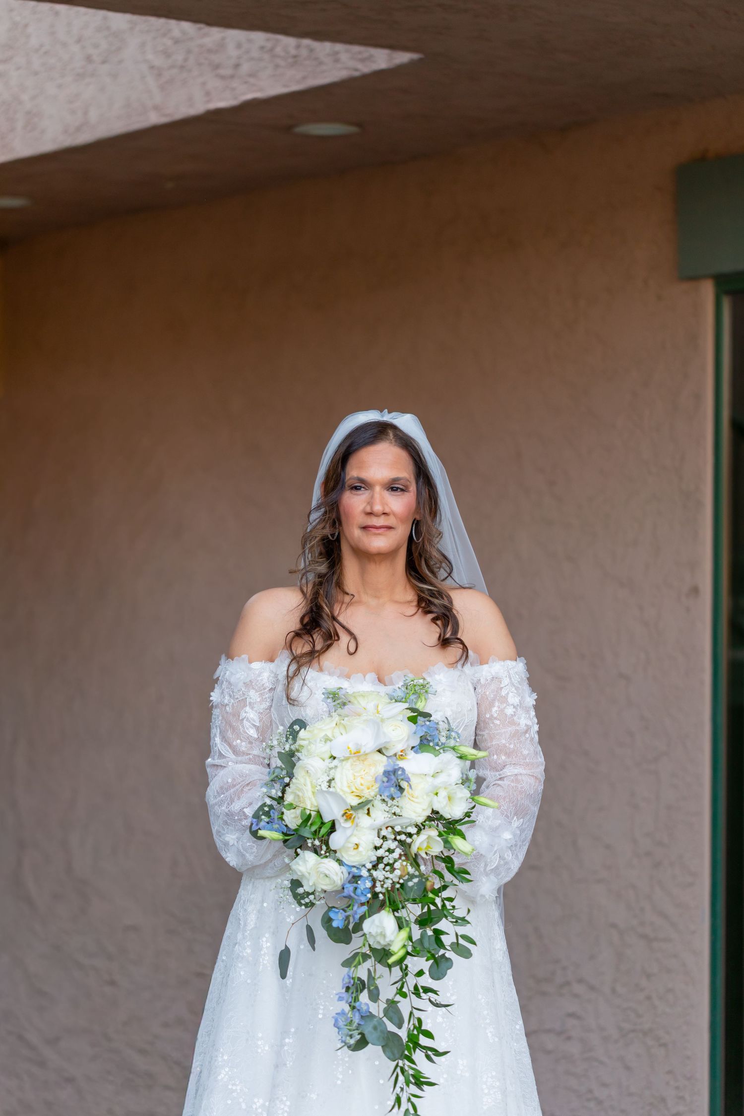 Bride in off-shoulder lace gown and blue veil holds cascading white and blue bouquet outdoors at Shugrues in Lake Havasu