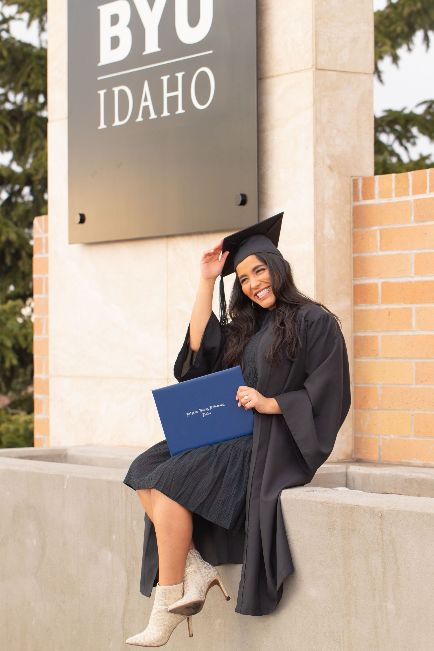 Smiling female graduate in black cap and gown holds diploma at BYU Idaho sign, wearing white boots.