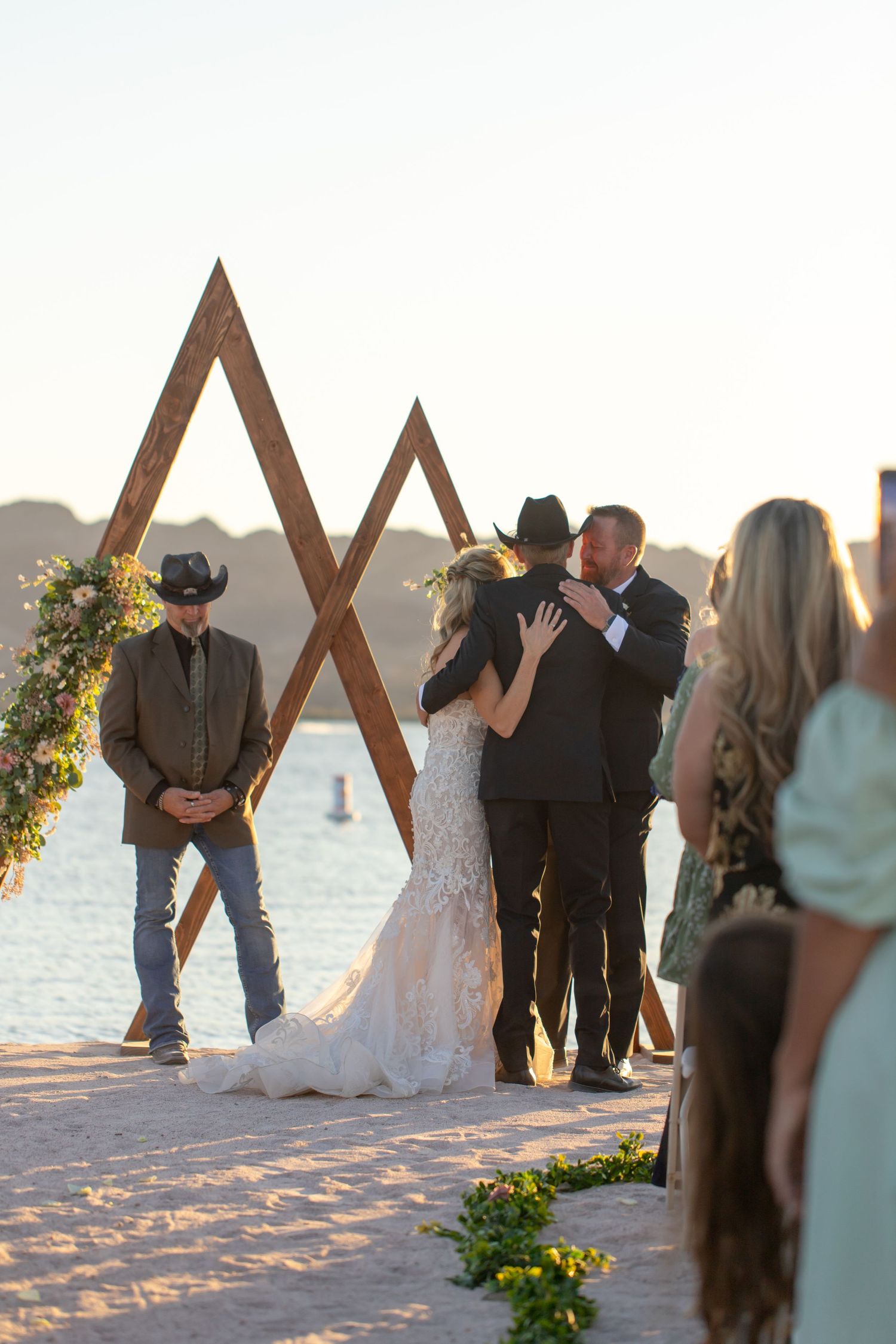 A wedding ceremony takes place at sunset under a wooden geometric arch decorated with flowers on a beach at the Riviera Marina in Havasu