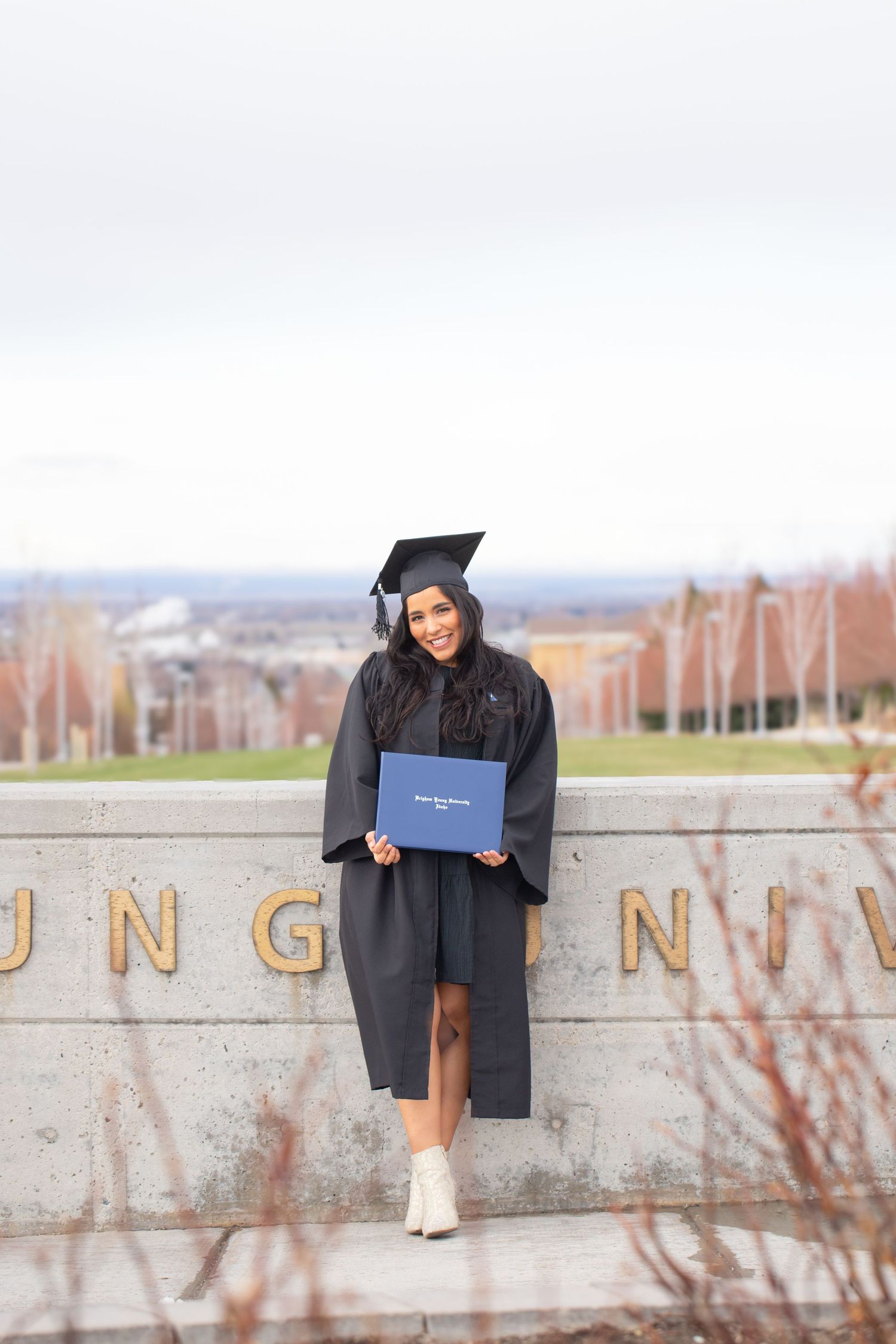 Young woman in black graduation cap and gown holding a blue diploma, smiling outdoors at a university campus.