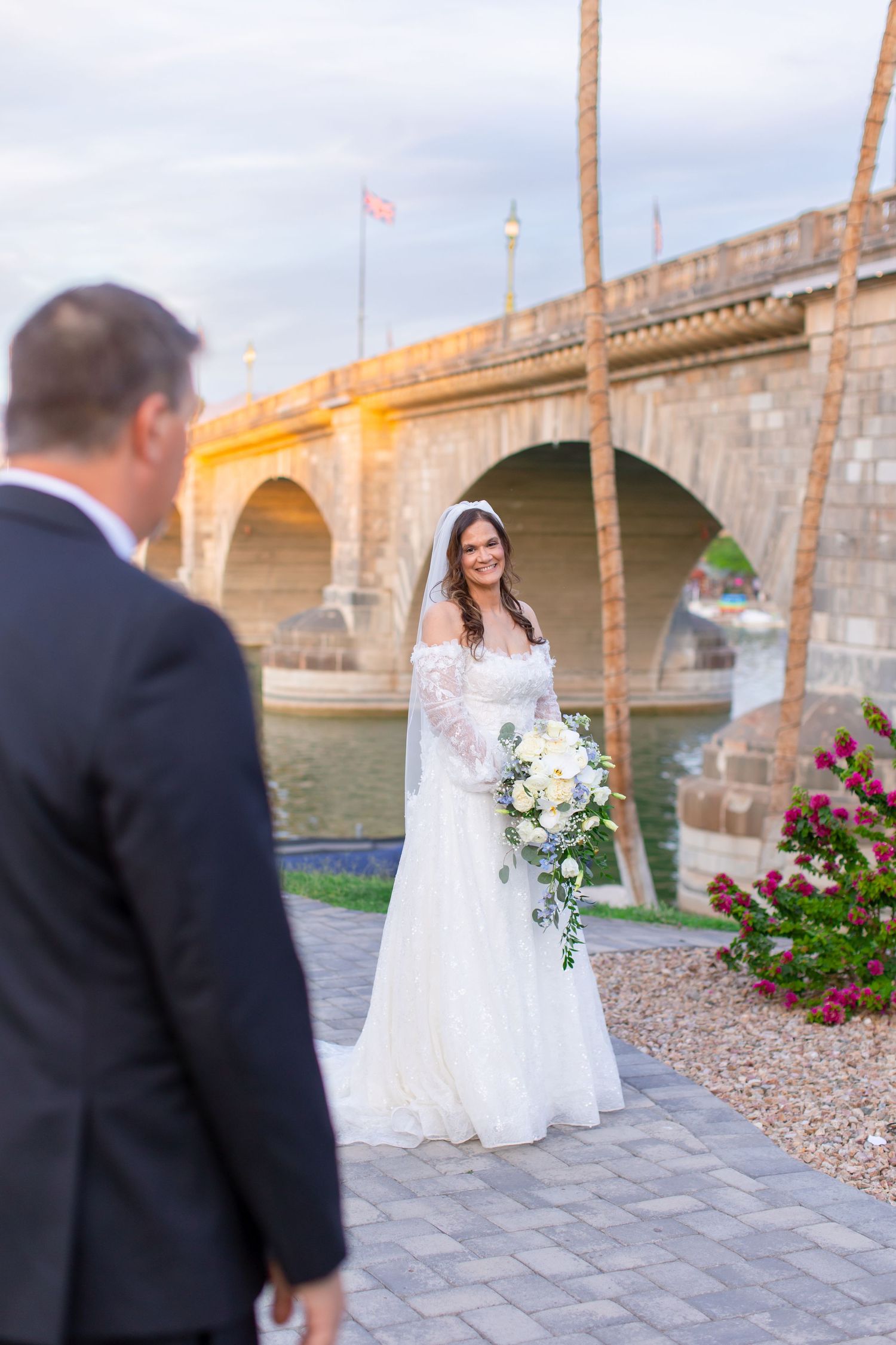 Bride in white lace gown holding bouquet smiles at groom near London Bridge in Lake Havasu City, AZ.