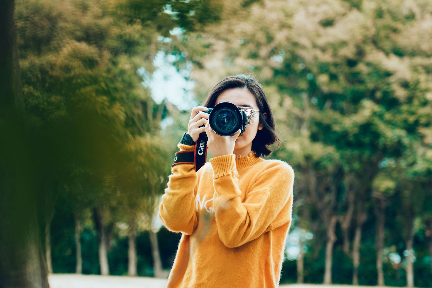 Photographer wearing yellow sweater taking pictures with professional camera in a wooded park setting.