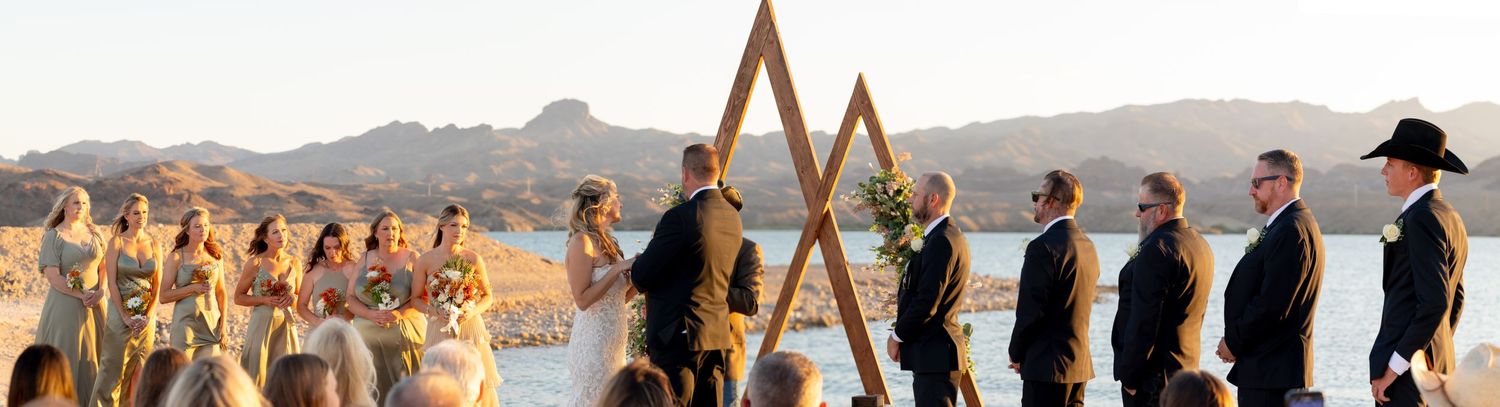 Wedding party stands in line near triangular wooden arch during golden hour in mountainous desert landscape at the riviera lake havasu