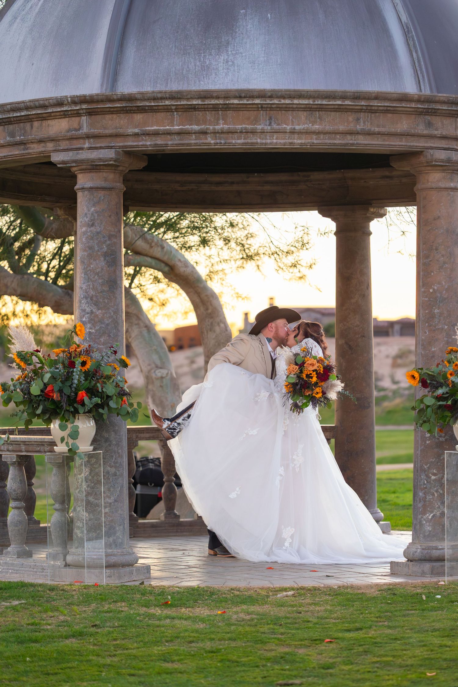 Couple kissing under iron wolf arch in lake havasu city arizona under stone colonnade with hanging floral arrangements at sunset.