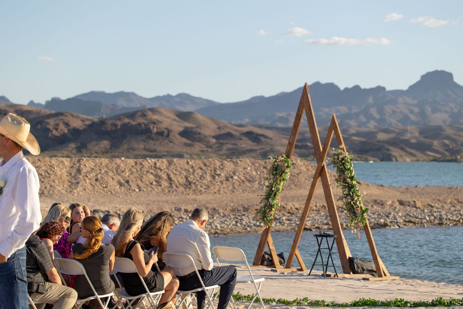 A scenic outdoor ceremony setup by a lake with mountains in the background, featuring a wooden triangle arch with greenery in Lake Havasu