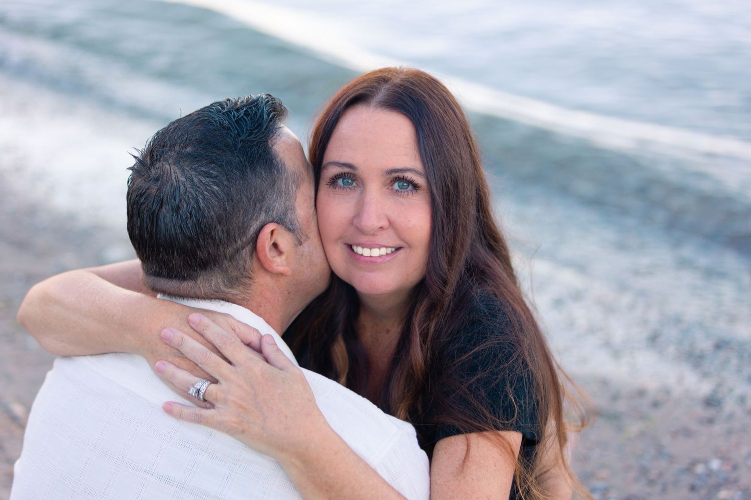 Smiling woman with long brown hair embraces a man in a white shirt on a beach, with soft ocean waves behind them.