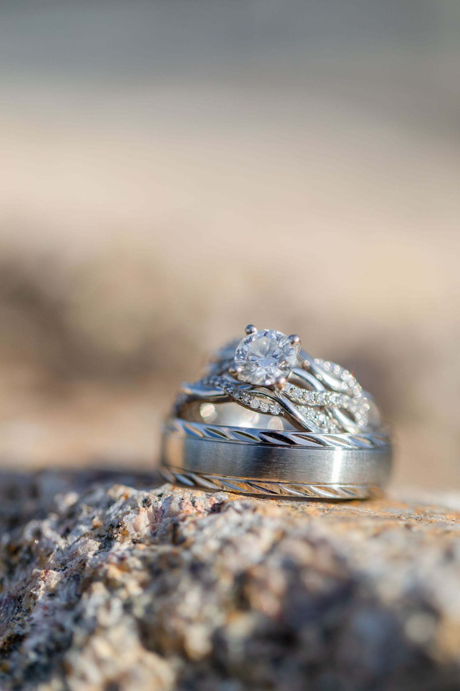Diamond engagement ring and wedding bands stacked on a rocky surface with soft bokeh background at Shugrues in Lake Havasu