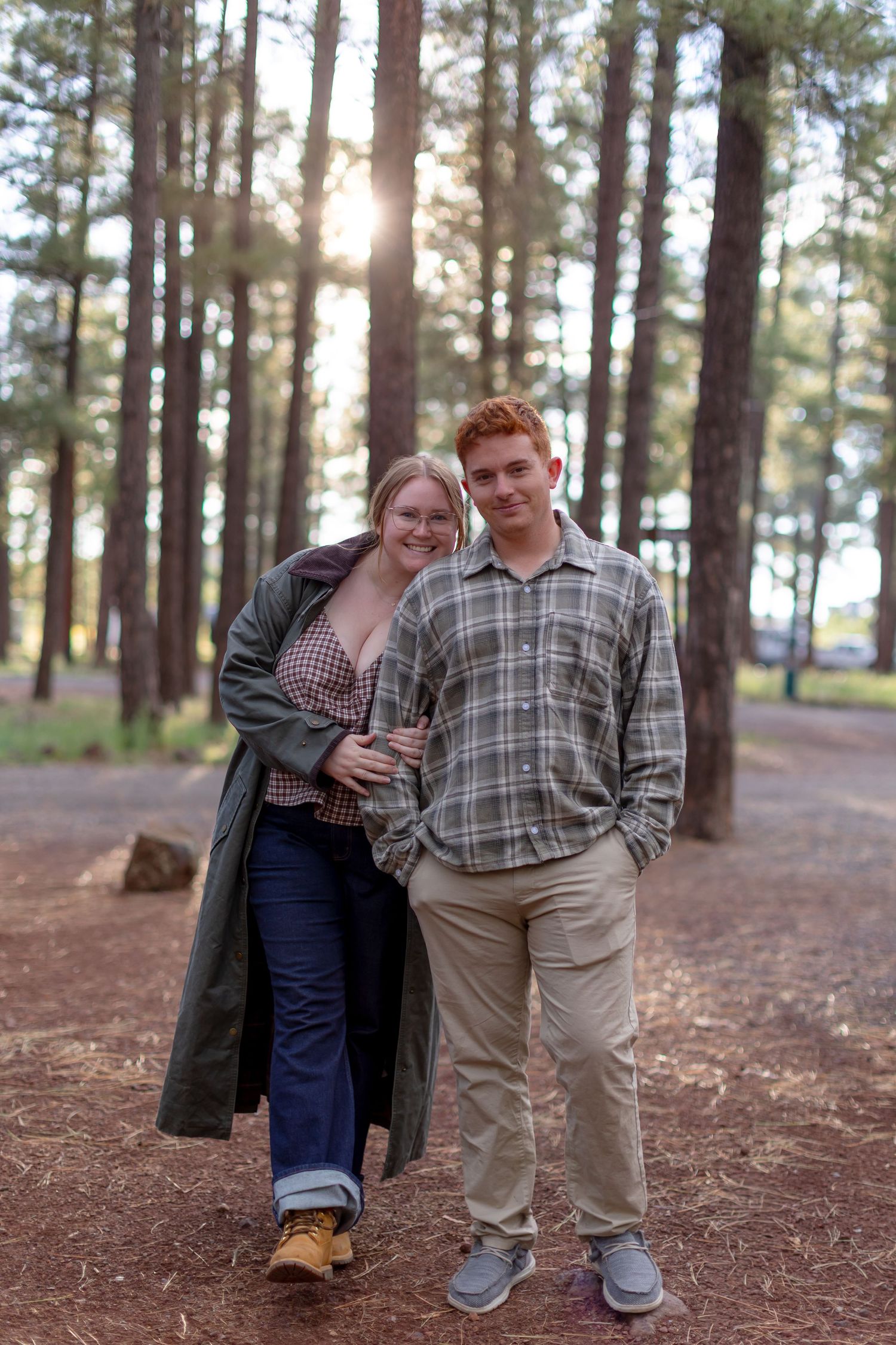 A young couple smiles together in a sunlit pine forest, woman in green coat and man in plaid shirt.