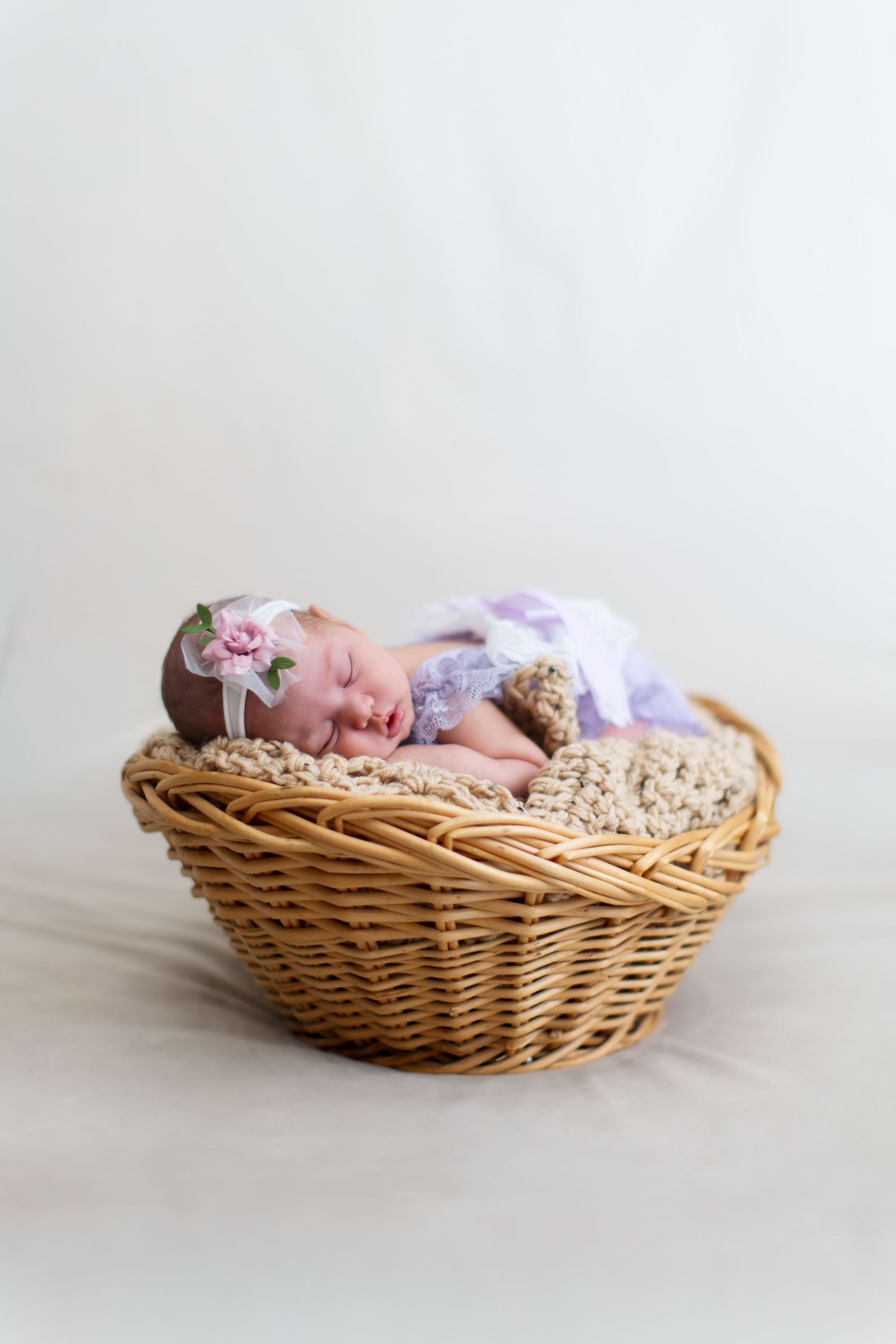 Newborn baby girl sleeping peacefully in a wicker basket, wearing a purple lace outfit and floral headband.