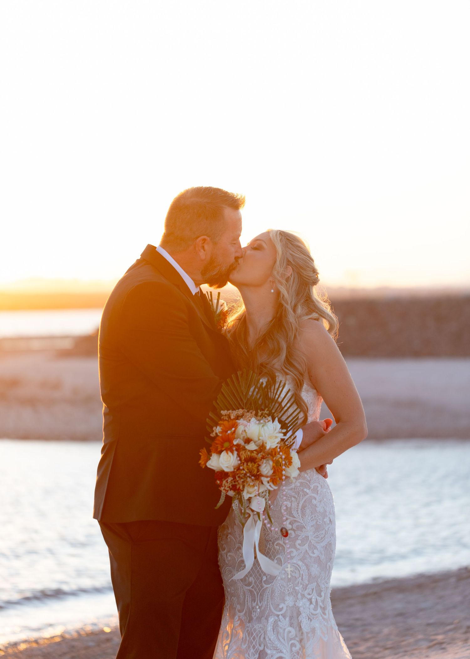 Couple kissing on beach at sunset, bride in lace dress holding bouquet at The Riviera Marina wedding.