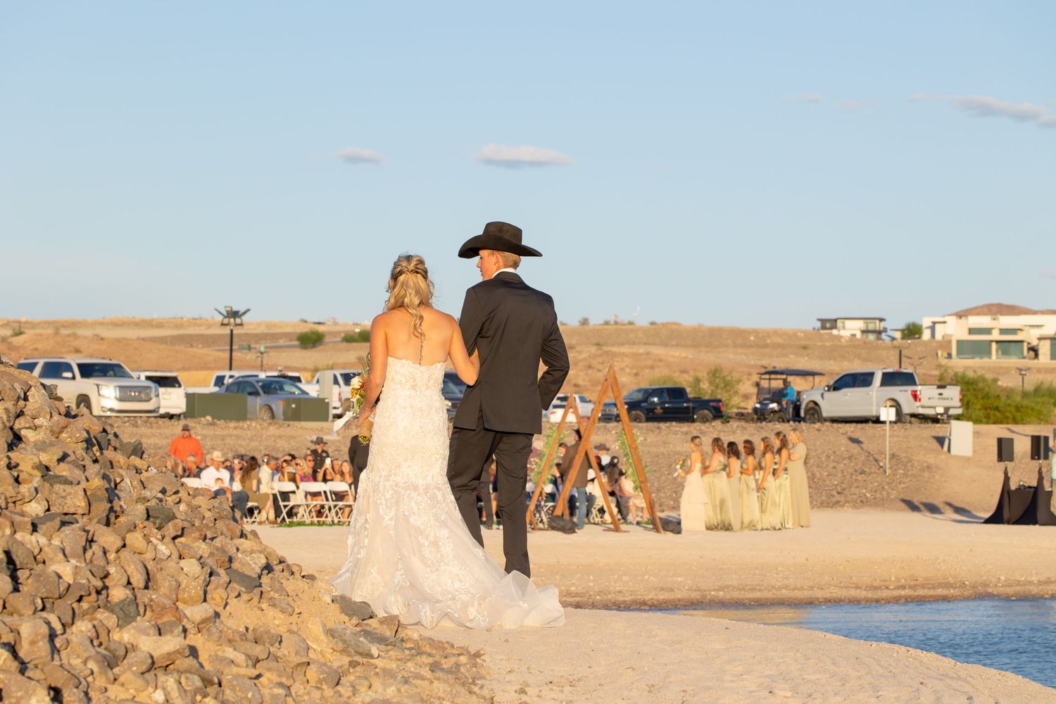 Couple in formal wedding attire walking together along a rocky coastal path during sunset with parked cars in background in Lake Havasu City