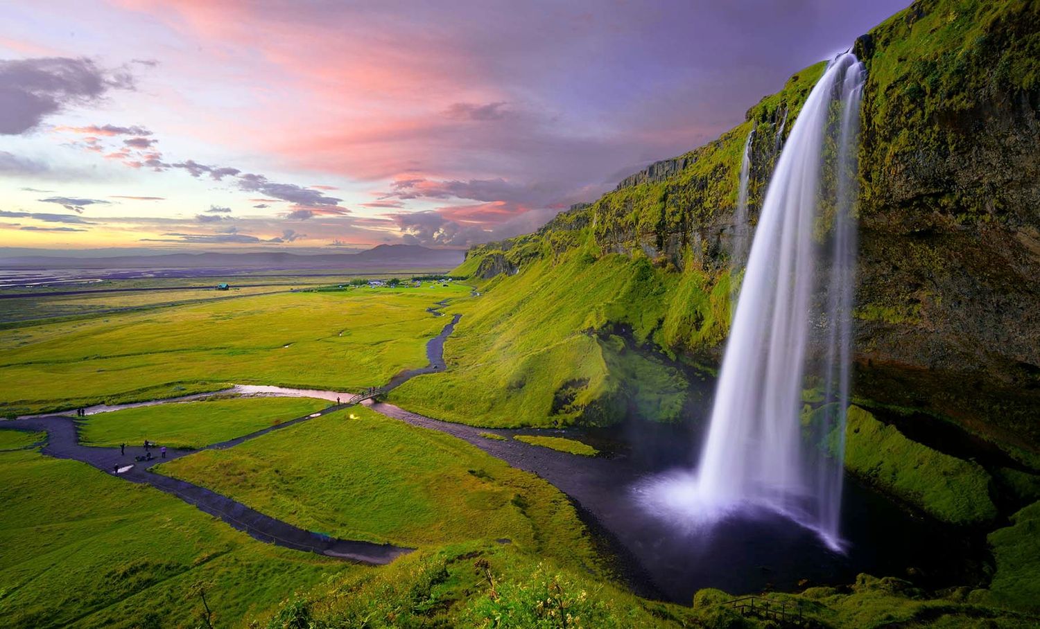 Majestic waterfall cascades down green cliffs at sunset in Iceland with winding river below.