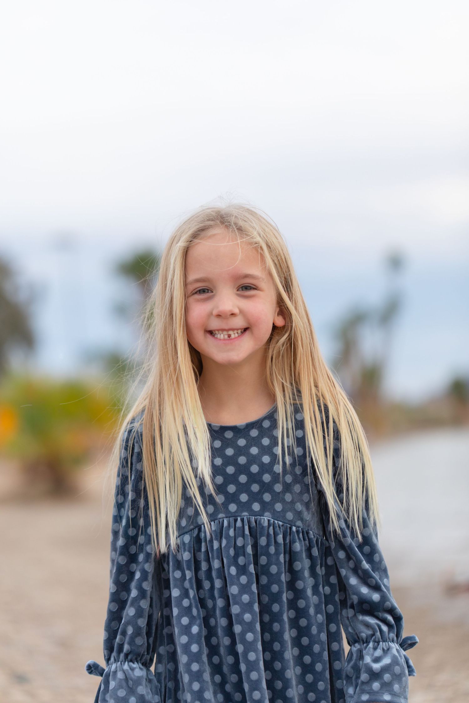 Smiling blonde girl with missing teeth wearing a blue polka dot dress outdoors near palm trees.