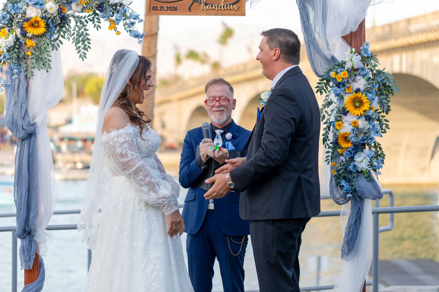 Couple exchanges vows at outdoor waterfront wedding ceremony with sunflower arch and officiant smiling.