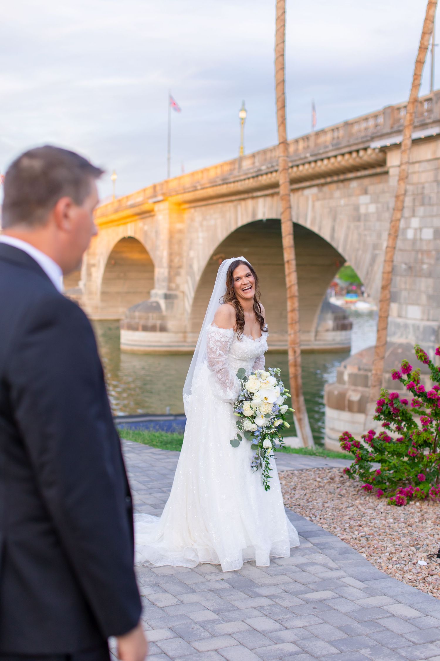 Bride in white gown holding bouquet smiles at groom near London Bridge in Lake Havasu City, AZ.